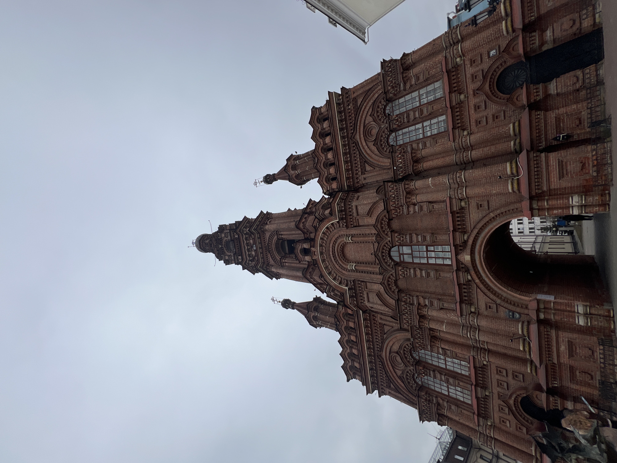 The image depicts a low-angle view of a building constructed of red brick, notable for its elaborate architectural details and soaring tower. The structure exhibits prominent arched windows, decorative moldings, and multiple smaller turrets. The sky is overcast, providing soft, diffused lighting.
Based on the distinctive architecture and the presence of a prominent tower, it appears to be the Kazan Family Center located in Kazan, Russia. The perspective emphasizes the building's height and ornate facade. The presence of an archway suggests it serves as a public space or entrance. The presence of people is slightly hinted in the lower right corner near the archway.