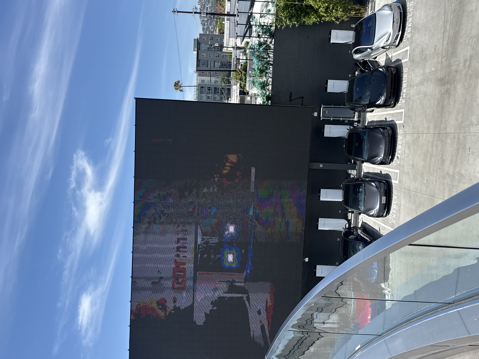 Captured from a high-angle perspective in West Hollywood, United States of America, the image displays a scene with a prominent outdoor digital display dominating the left side of the frame. The screen shows a distorted image.
To the right of the screen is a wall, alongside which are Tesla charging stations with several Tesla vehicles parked. The Teslas appear to be charging. The parking spaces are marked with "CHARGING ENABLED". A palm tree, several mid-rise buildings, and electric power lines are visible in the background. The sky is clear and blue. Part of a modern building's architectural detail, featuring curved lines, is in the lower-right foreground.