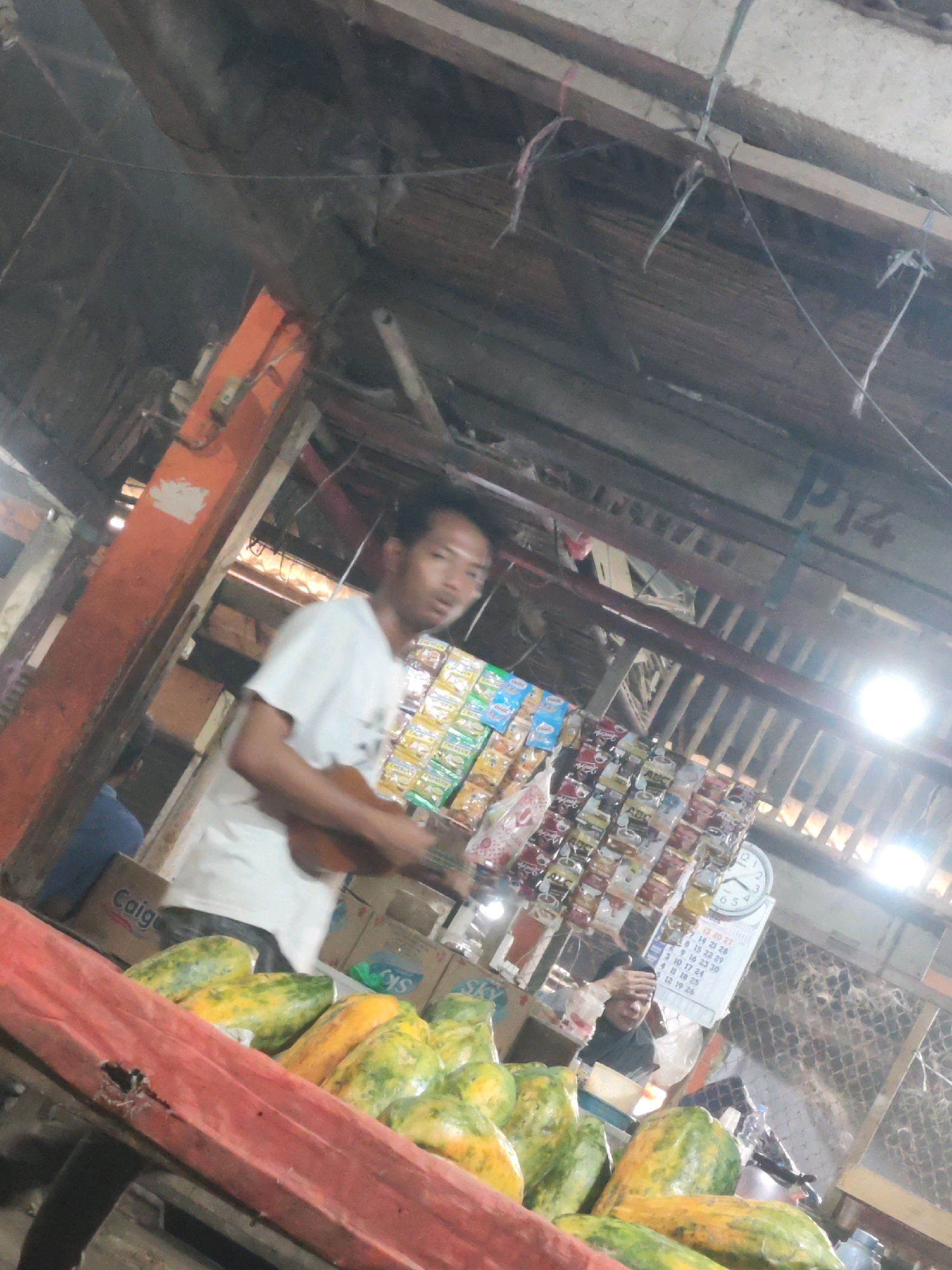 The image depicts a man playing a ukulele in what appears to be an outdoor market stall in Karawang, Indonesia. The stall has a rustic wooden roof structure with exposed beams and wires. In the foreground, a red-covered cart displays a pile of papayas. Behind the man, a variety of packaged snacks and goods are displayed on shelves. A clock and a calendar are visible on the wall behind the stall. The overall lighting suggests it is daytime, with a bright light source possibly a lamp or sunlight visible on the right side of the frame. The man is wearing a white t-shirt and is the central human subject in the image.