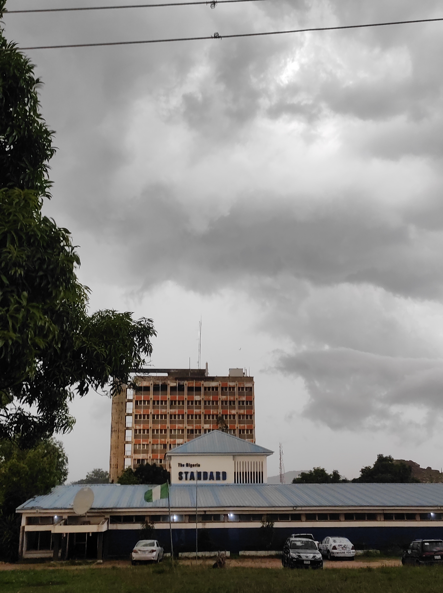 The photograph captures a scene in Jos, Nigeria, featuring the office of The Nigeria Standard newspaper. A partially visible tall, weathered building stands behind the single-story newspaper office, which has a blue roof and a sign displaying the paper's name. The Nigerian flag is seen near the building. Several cars are parked in front of the newspaper office. The sky is overcast. A tree with green leaves is visible in the foreground.