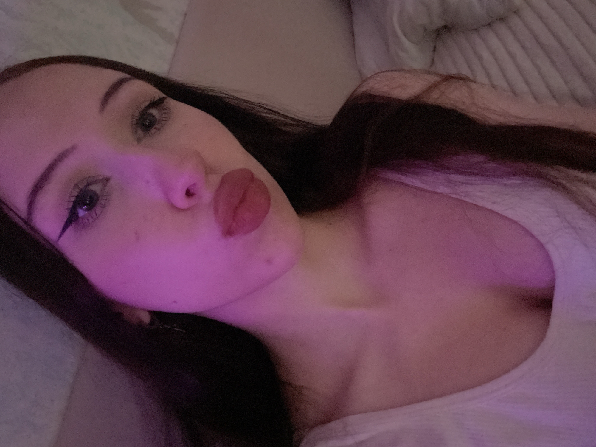 The image is a close-up selfie of a young woman with dark hair. She is wearing a light-colored tank top and has makeup on, including winged eyeliner and lipstick. The lighting in the image is primarily pink, casting a purple hue on her face and upper body. The background appears to be a bed with striped bedding. There is no information available to determine the location of this image in Votkinsk, Russia.