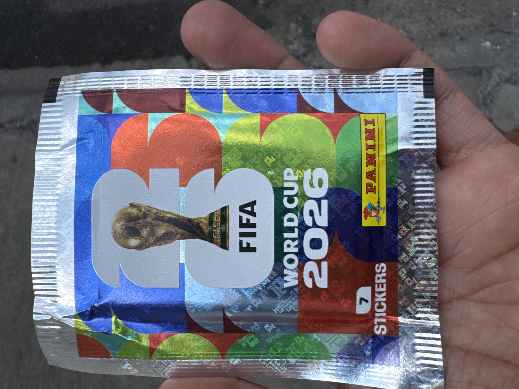 A person's hand holds a sealed package of FIFA World Cup 2026 Panini stickers. The package is shiny and features the FIFA World Cup trophy, "FIFA World Cup 2026," and "7 STICKERS" text. The package has various colorful geometric designs as a background. The background is slightly out of focus, but suggests a street or sidewalk with a gray concrete surface. Based on the available information, the image was taken in Puebla, Mexico.