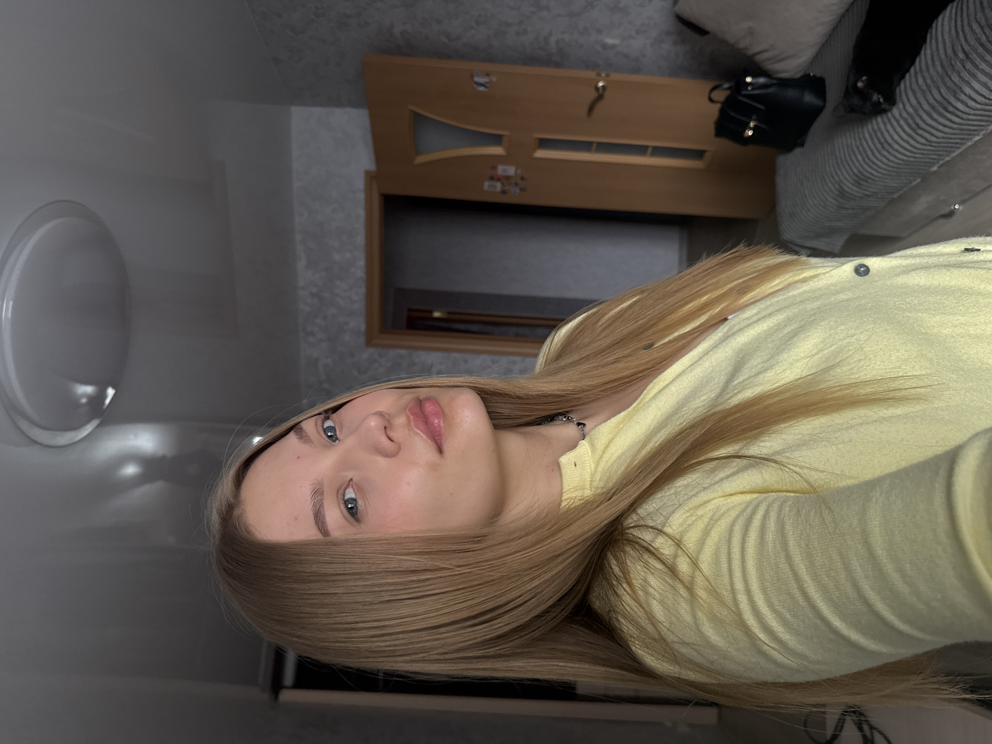 The image is a selfie of a young woman with long blonde hair, wearing a light yellow top. She has fair skin, blue eyes, and is looking at the camera with a neutral expression. The background features a doorway leading to a room with a sofa, a black purse or bag, and a wall with a light fixture. The location cannot be identified as Chaikovsky, Russia, with the given information.