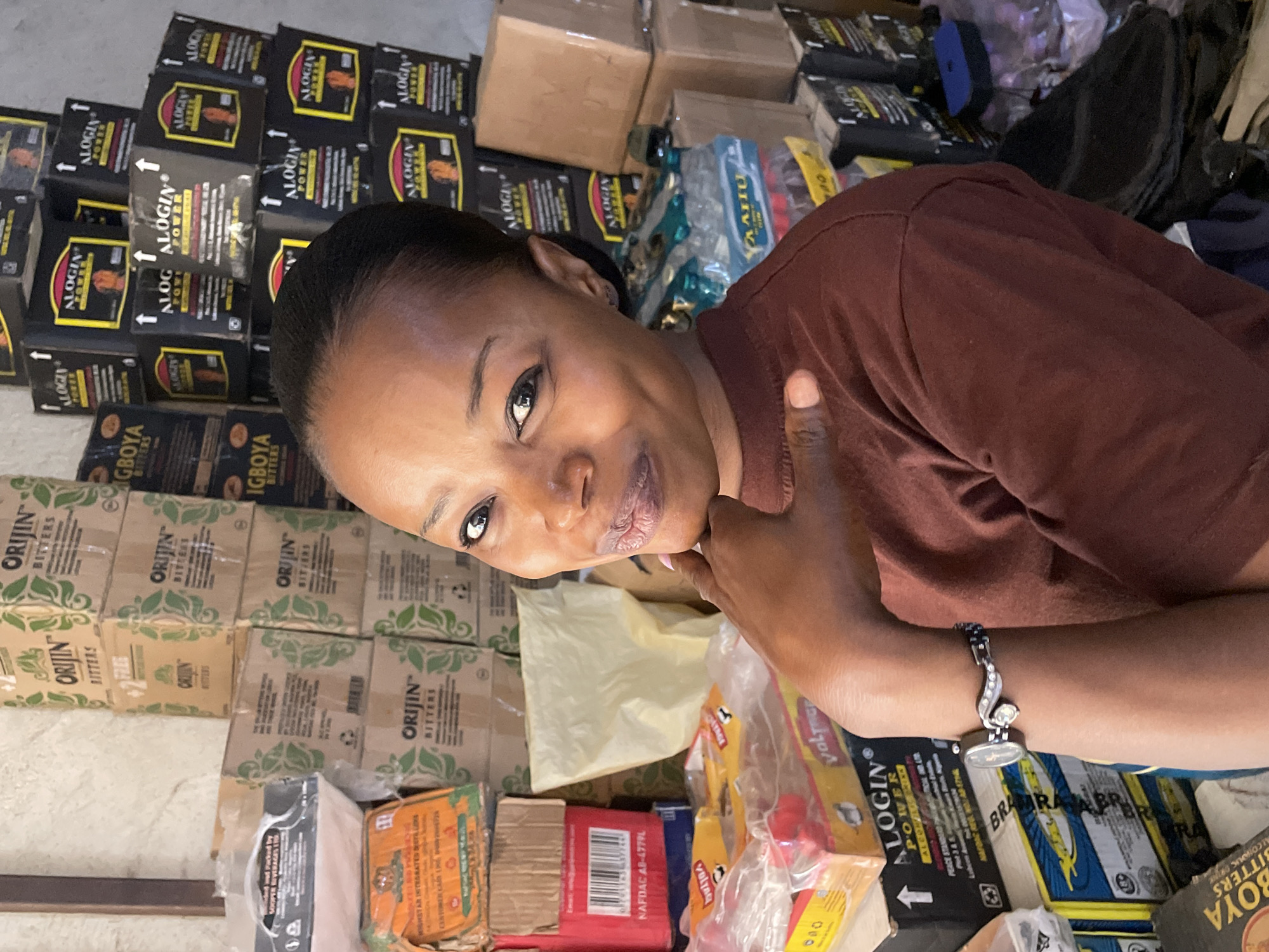 The image shows a woman with dark hair, wearing a dark red shirt, smiling and giving a thumbs-up. She has a friendly expression and is wearing a decorative silver-colored watch on her left wrist.
The setting appears to be an indoor commercial space, likely a shop, market stall, or storage area, in Jos, Nigeria. The background is densely packed with various packaged products, neatly stacked in boxes and bags.
Notable products visible include numerous black boxes labeled "ALOGIN POWER," often featuring an upward arrow symbol. Many cardboard boxes with green leaf designs are stacked, clearly showing the brand "ORIJIN BITTERS." Other boxes are identifiable by text such as "IGBOYA BITTERS" and "BRARABA." Smaller clear plastic bags contain items labeled "VOLTAGE." Regulatory information, "NAFDAC AB-4779L," is visible on one red box. The woman's positive gesture and the abundance of products suggest she might be a proprietor, vendor, or staff member showcasing the goods in a bustling commercial environment. The image is taken at an oblique angle.