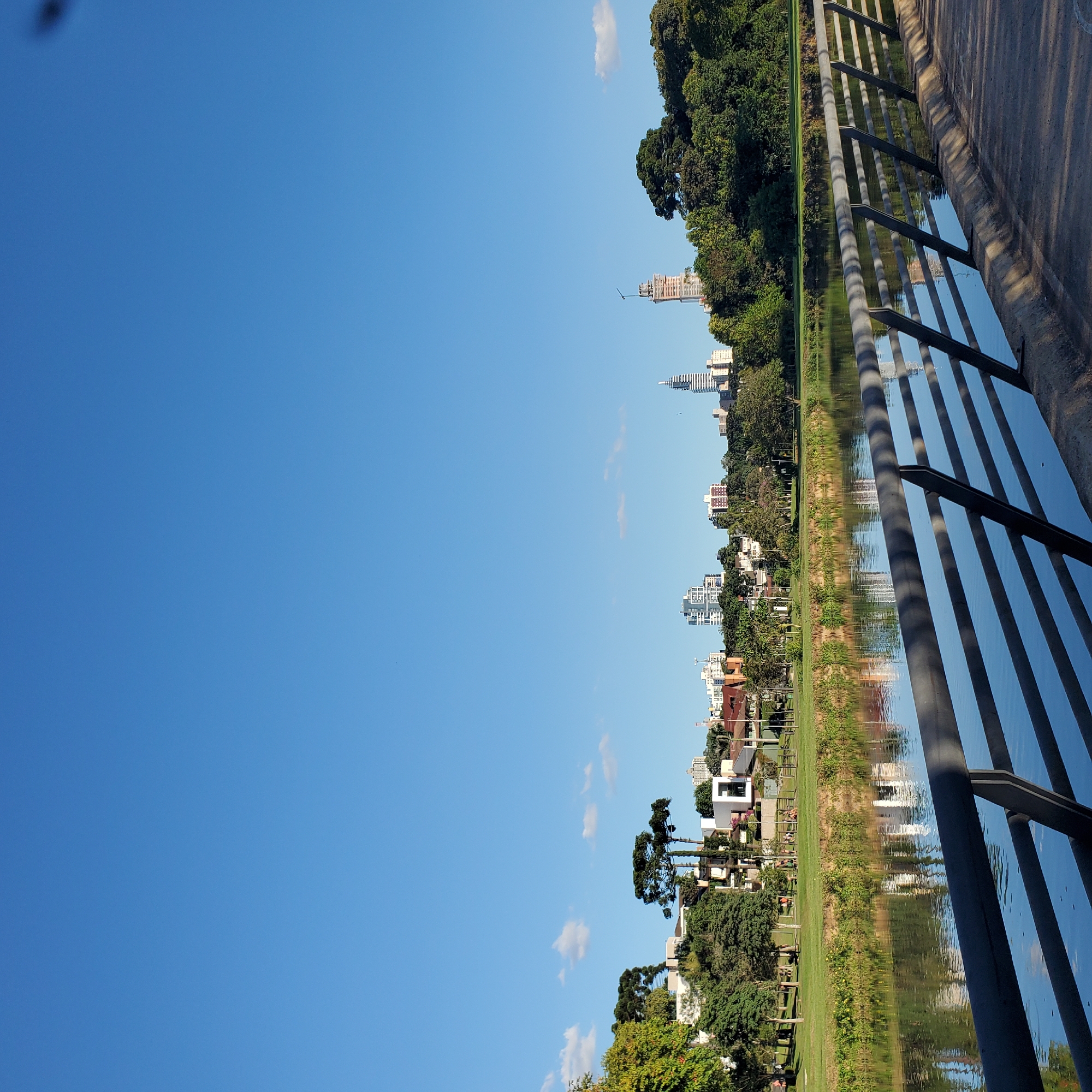 A serene, wide-angle, outdoor daytime shot reveals a section of the Iguaçu River within the city of Curitiba, Brazil. The clear blue sky, occupying a substantial portion of the image, contrasts with the vibrant green landscape along the riverbank. A mix of lush trees, manicured lawns, and residential buildings is visible. In the distance, the city skyline includes high-rise buildings. The water's surface reflects the sky, vegetation, and urban structures. A metallic railing of a bridge or walkway is visible on the right.