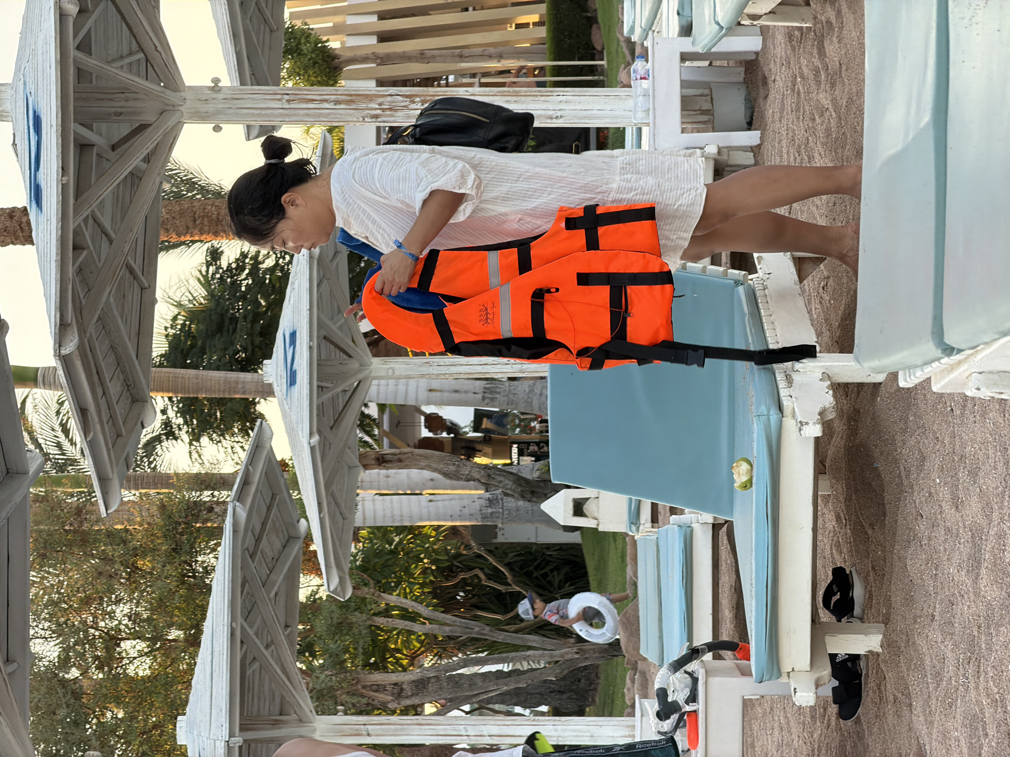 A woman stands on a sandy beach, holding an orange life vest. She is wearing a white dress and a black backpack. In the background, there are several white beach umbrellas and lounge chairs. A young boy walks in the distance holding a white inflatable ring. The location is a resort in Sharm El Sheikh, Egypt, with palm trees and other tropical foliage visible. The lighting suggests it is either early morning or late afternoon.