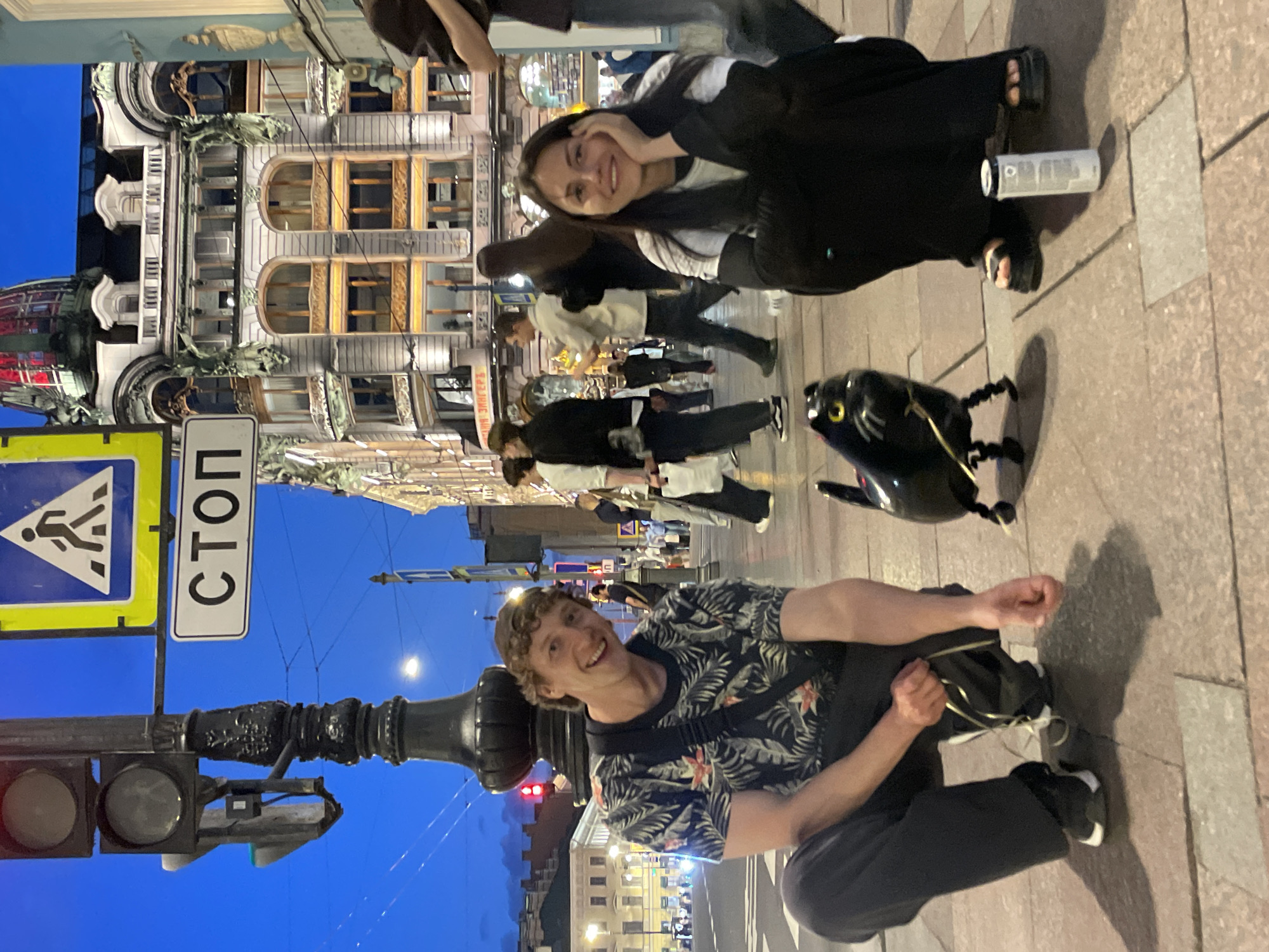 The image depicts an evening street scene on a paved sidewalk in Saint Petersburg, Russia, with the Singer House (Dom Knigi) prominently featured in the background. Two individuals are squatting in the foreground, smiling directly at the camera. On the left, a man with curly brown hair, wearing a dark tropical-patterned t-shirt and dark pants, holds the string of a small, black, panther-shaped walking balloon with yellow eyes. On the right, a woman with long dark hair, wearing a dark jacket over a white long-sleeved top and dark pants, rests her chin on her hand. A white cylindrical object lies on the ground beside her. A black, ornate lamppost stands in the mid-ground, supporting a traffic light, a standard pedestrian crossing sign (blue triangle with a white figure on a yellow square background), and a white rectangular sign displaying "СТОП" (STOP) in black Cyrillic letters. The Singer House, an elaborate multi-story building with a distinctive red-tinted glass dome and numerous ornate windows, is brightly illuminated in the background. Several blurred pedestrians are visible walking on the street and sidewalk. The sky is a deep blue, indicating twilight or early night.