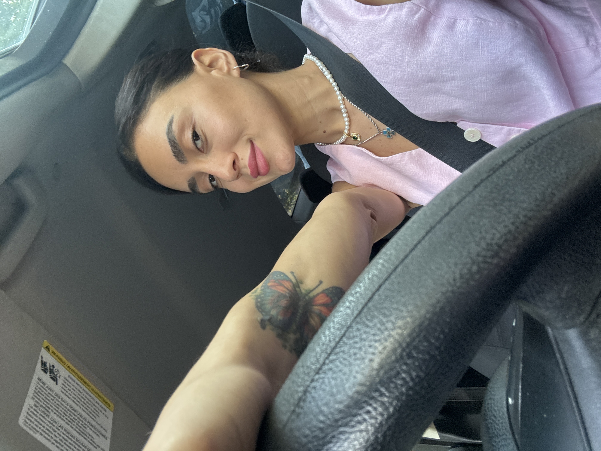 A woman with dark hair pulled back is seen from the chest up, inside a car. She is wearing a pink button-up shirt and a pearl necklace with a silver pendant. Her seatbelt is fastened across her chest. Her left arm is resting on the steering wheel, and a butterfly tattoo is visible on her forearm. She is looking towards the camera with a slight smile. The interior of the car is visible, including the steering wheel and a sun visor with warning labels. The scene appears to be set during the daytime, with natural light entering the car.