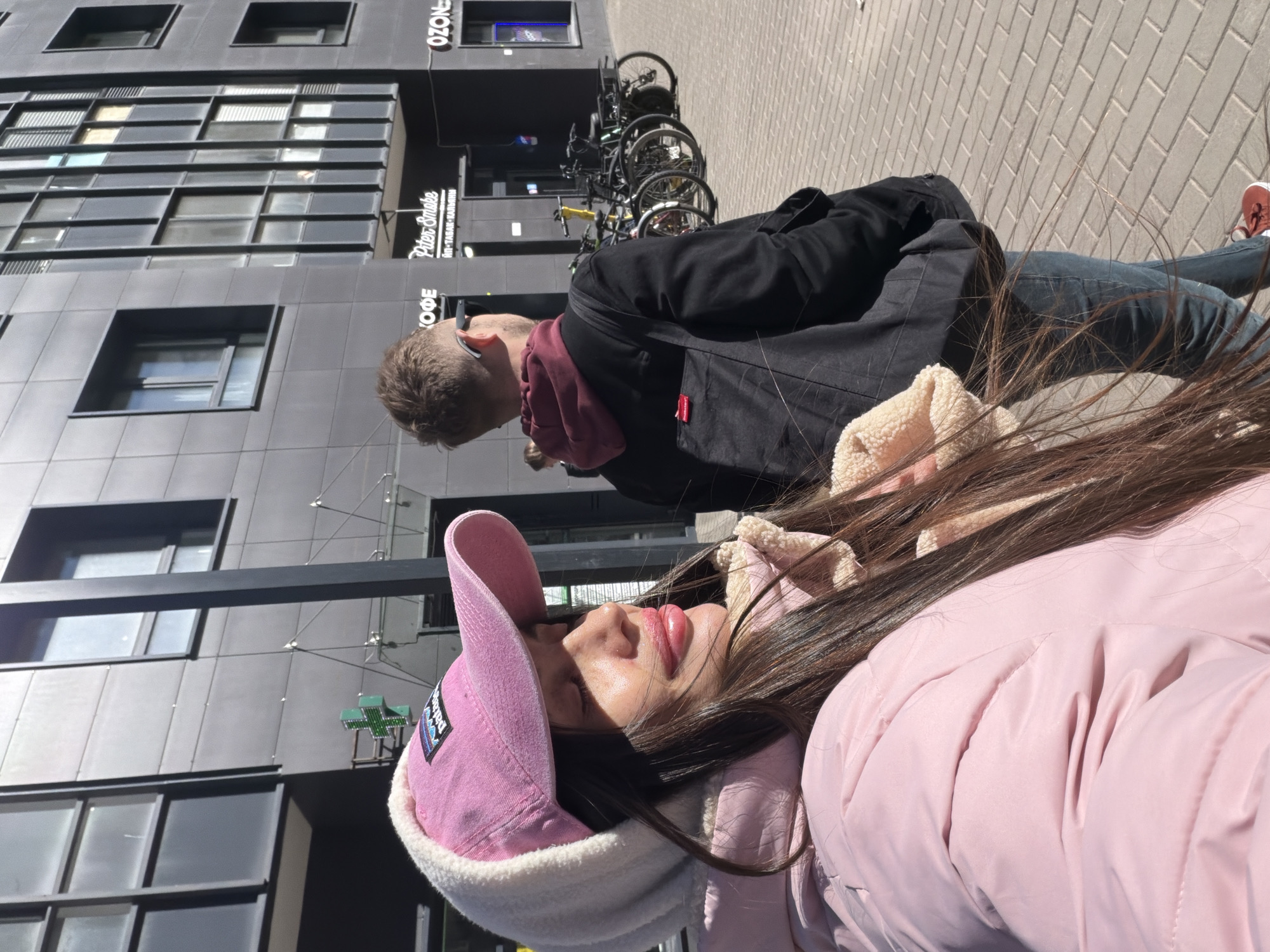 The photograph shows two individuals on a street. A woman with long, dark hair is in the foreground, wearing a pink jacket and a pink cap with a white fur trim. Her eyes are closed, and she appears to be lying down. Behind her, a man with short hair, wearing a dark jacket and a maroon scarf, stands on the sidewalk. Bicycles are parked near the building behind the man. The building is gray with dark-framed windows, and signs for "Peter Smoke" and "OZON" are visible. The street is paved with light-colored bricks. The scene is set in Saint Petersburg, Russia, as evidenced by the architectural style and the Cyrillic script on the signs.