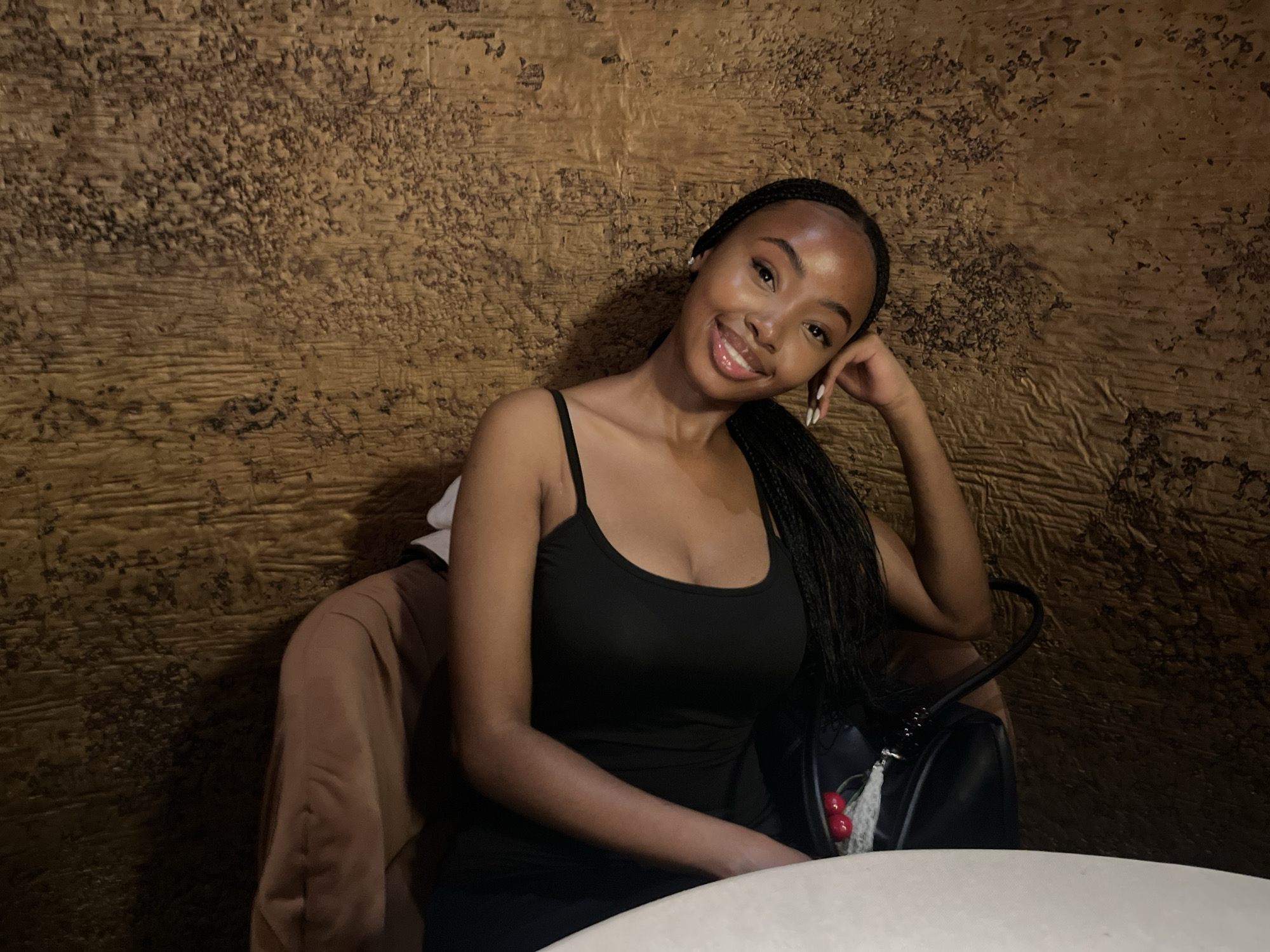 A young Black woman with braided hair and a black tank top smiles as she rests her head on her hand. She is seated in a booth with a textured, gold-colored wall behind her. A brown jacket is draped over the back of her chair, and a black purse with a cherry charm is visible next to her. A white table is in the foreground. The location is not specified as Kazan, Russia.
