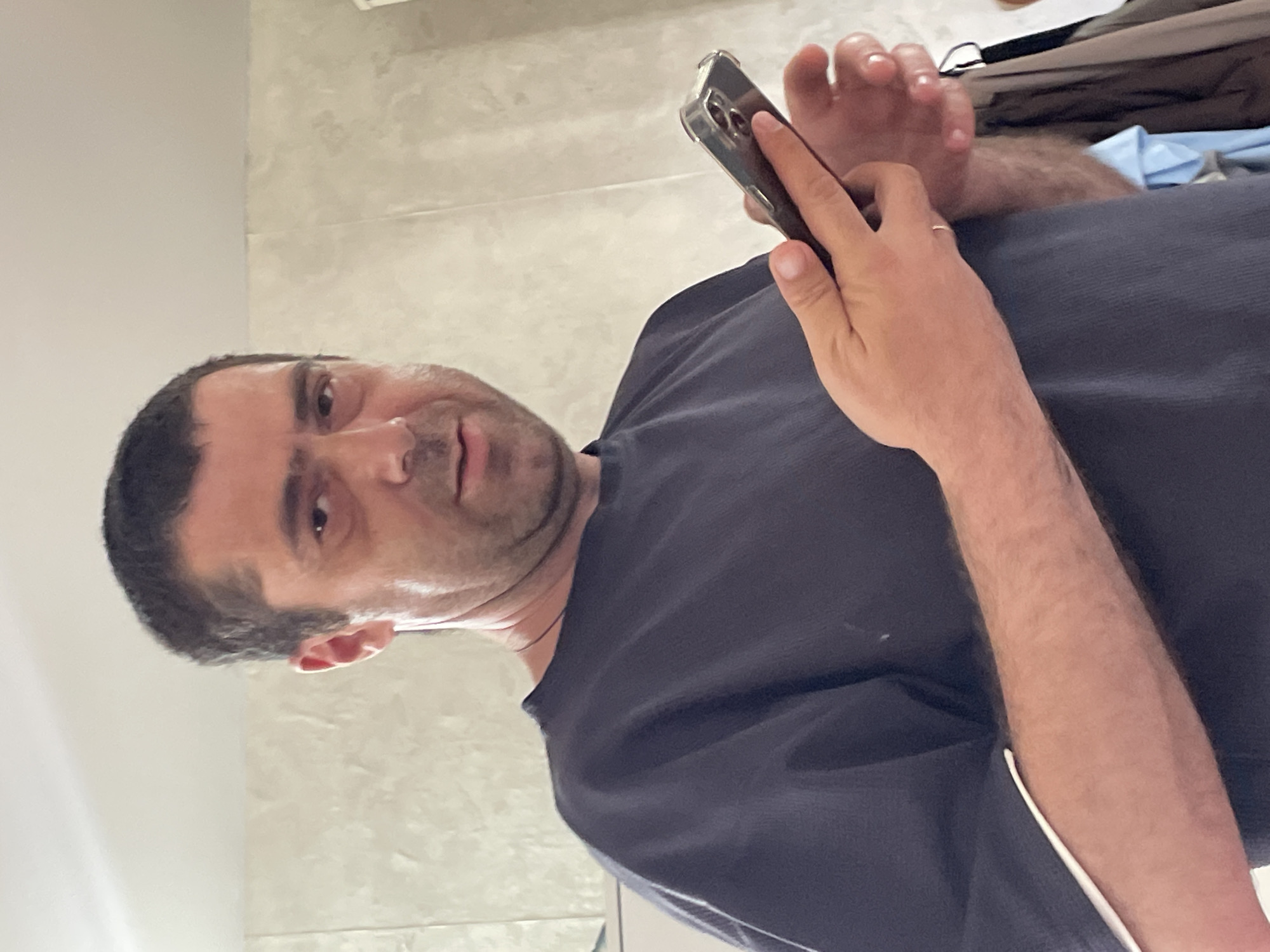 A man with short, dark hair and a receding hairline, wearing a dark blue t-shirt with white trim on the sleeves, is holding a smartphone in his right hand and looking at the camera. He has a serious expression and appears to be in an indoor environment with a textured, light-colored wall behind him. The scene suggests a casual, everyday moment.