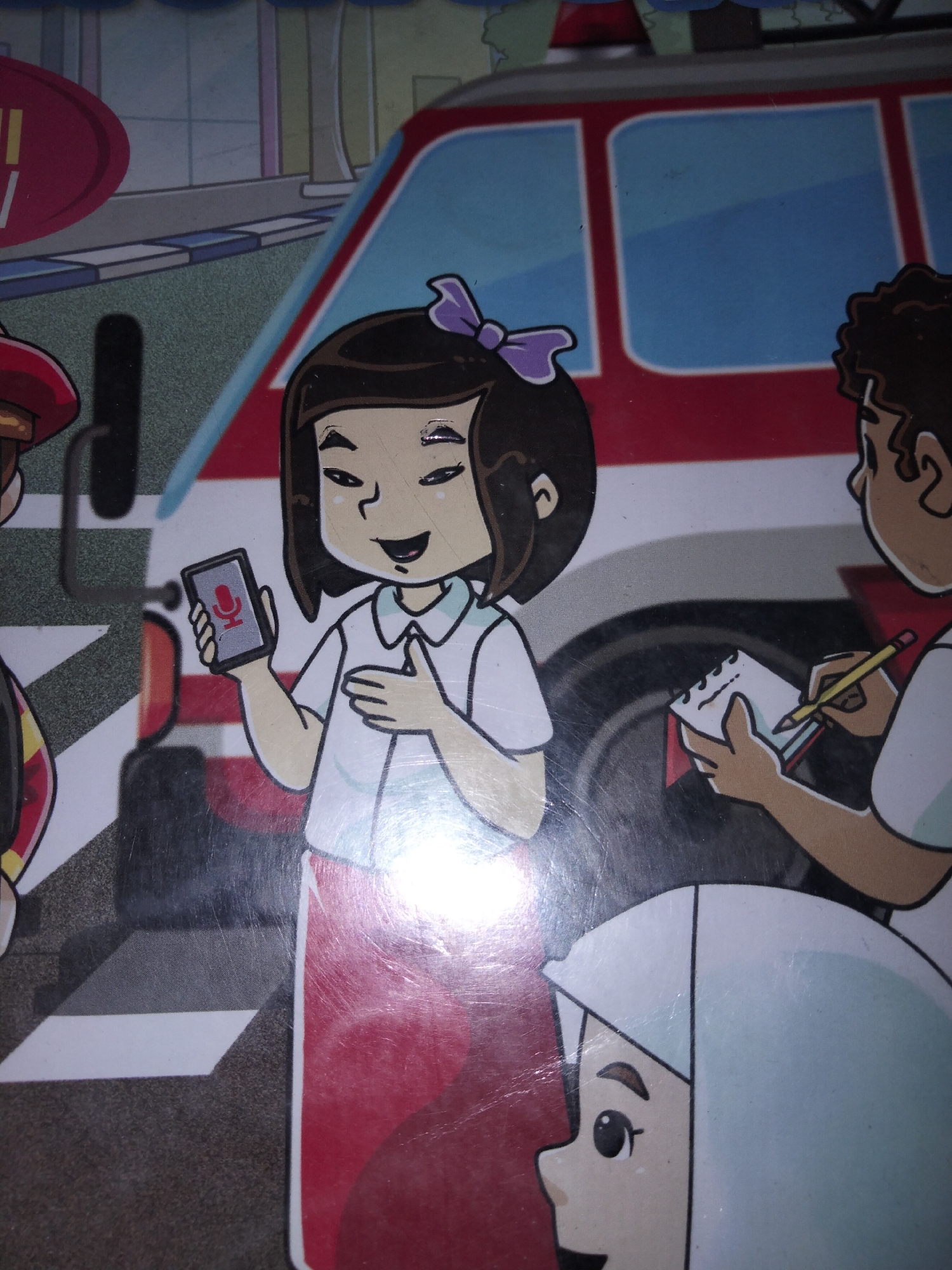 In a brightly colored, cartoonish depiction of an urban scene, likely within the fictional city of Payagaboh, Indonesia, a group of individuals are gathered near a red and blue fire truck. The foreground features a young girl with dark hair pulled back by a purple bow, wearing a white shirt and a red skirt. She is holding up a smartphone displaying a red microphone icon, suggesting she is either recording or participating in a call. Her expression is cheerful. To her right, another person, possibly a reporter, is shown from the shoulders up, wearing a red collar and a white shirt. They are actively writing on a small notepad with a yellow pencil, their gaze directed downwards at the pad. Partially visible to the left is another figure in a red helmet, possibly a firefighter, and at the bottom right, the side profile of a person wearing a light blue head covering is visible. The background includes elements of a city street with a building facade and sidewalk. The overall impression is one of civic engagement or a news report related to emergency services.