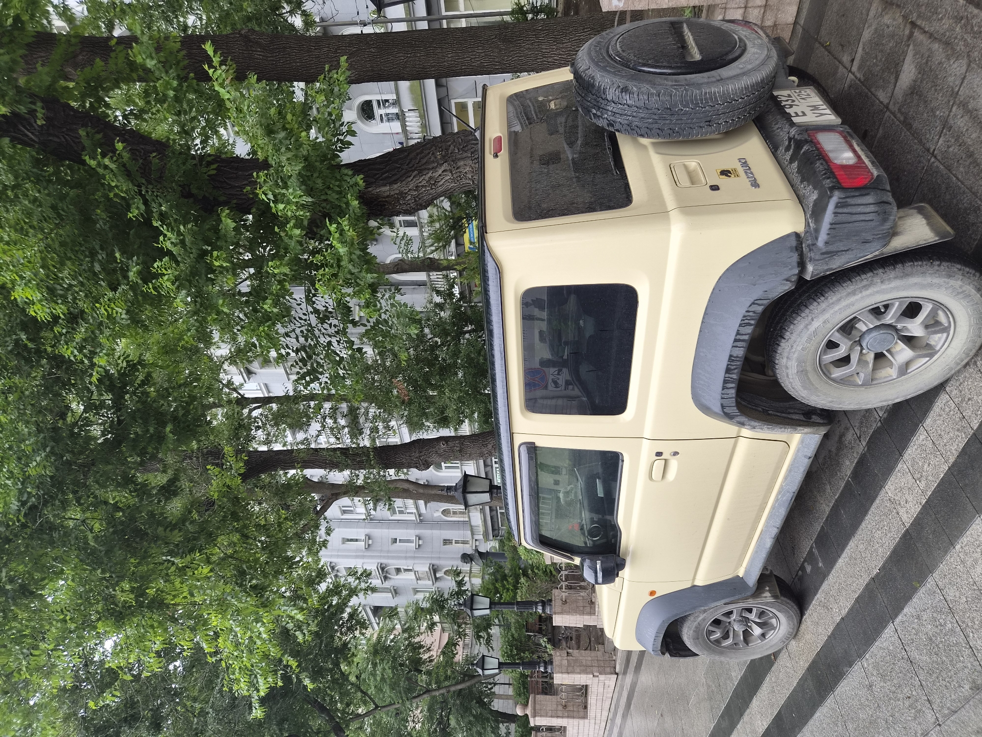 The image shows a light yellow Suzuki Jimny parked on a wet, paved walkway in Vladivostok, Russia. The car is facing away from the camera, with its rear bumper, spare tire, and driver's side visible. The walkway is composed of alternating dark gray and light gray rectangular paving stones arranged in diagonal stripes. Behind the car, a building with multiple stories and arched windows can be seen. Several large trees with green foliage are present in the foreground and background, obscuring parts of the building. Streetlights and a bust on a pedestal are also visible to the left of the car, suggesting a public space or park area. The overall impression is that of an overcast day, with moisture on the car and pavement. The license plate of the Suzuki Jimny reads "E 932 YM 125".