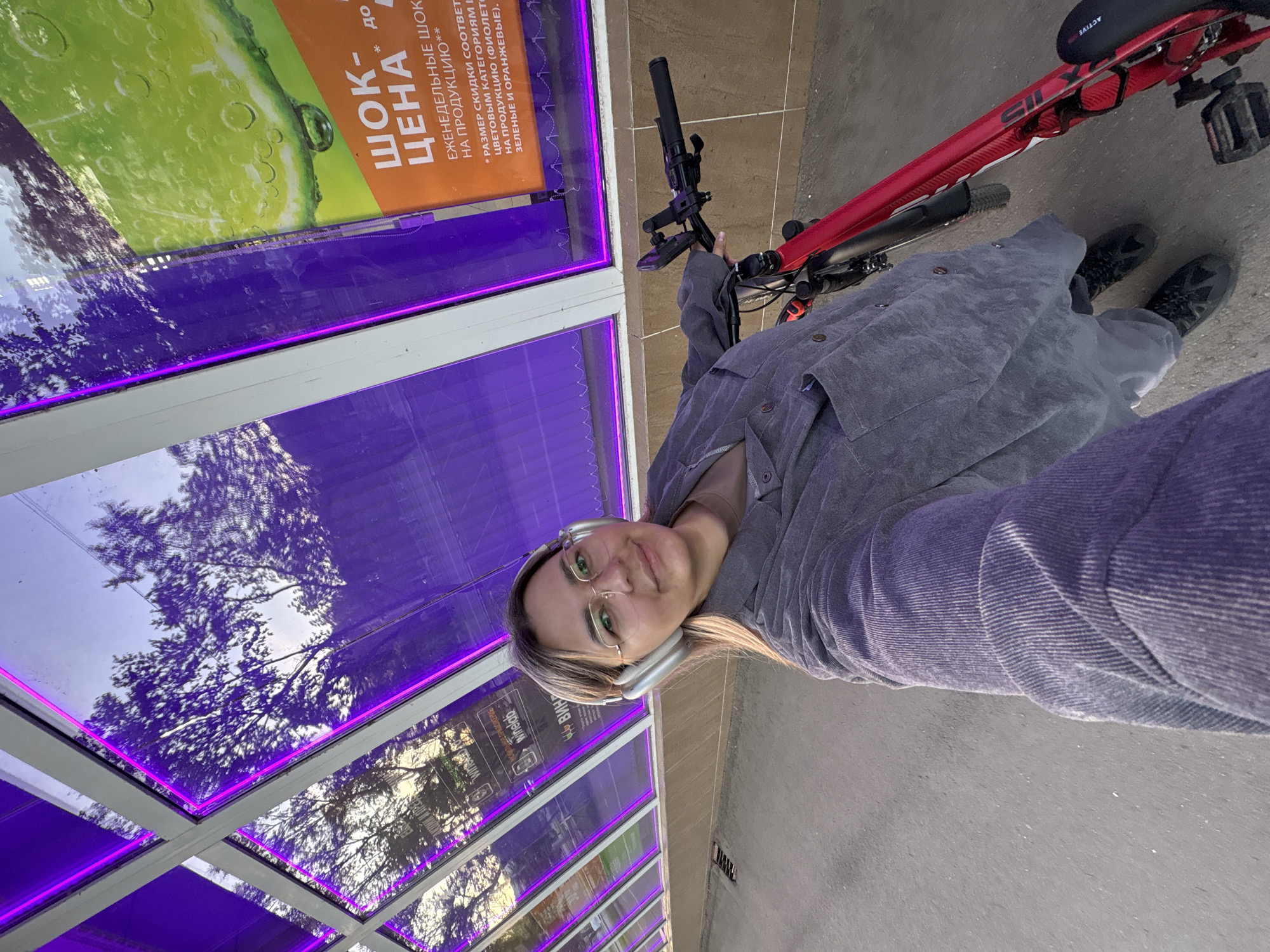 A woman with blonde hair and glasses poses for a selfie with a red bicycle next to a storefront in what is most likely Birobidzhan, Russia. She wears a gray button-down shirt and headphones, and is looking towards the camera. The bicycle is partially visible, positioned to her right. The storefront features windows with purple neon lighting and advertising posters in Russian, including a promotion with the word "Шок-Цена" (Shock Price). Reflections of trees are visible in the glass. The location appears to be a covered walkway or bus stop shelter with concrete ground.