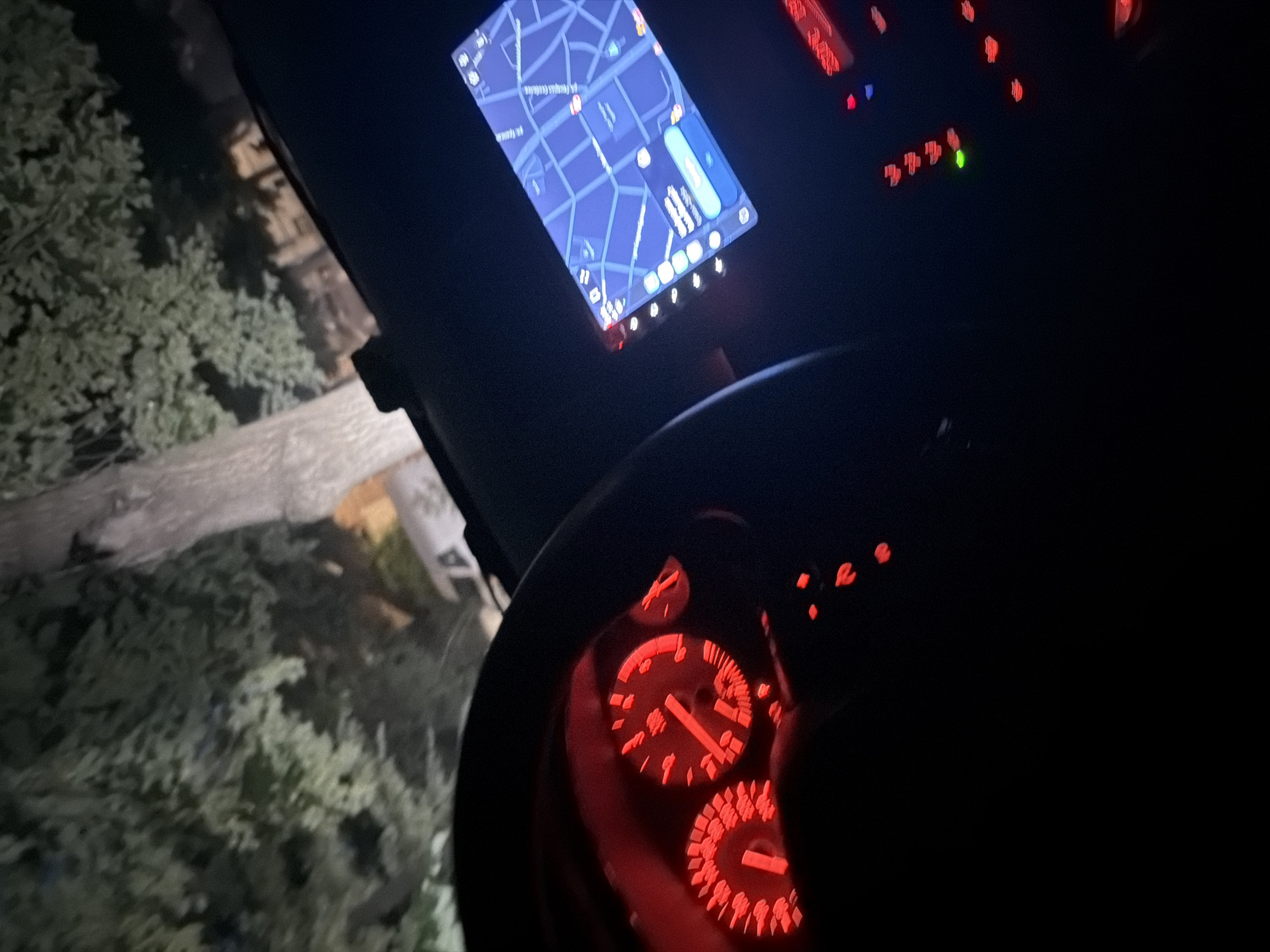 The image shows the interior of a car at night, with the dashboard illuminated by red lights from the instrument cluster and a navigation screen displaying a map. The exterior of the car shows trees and a streetlight, suggesting it is parked on a street in a city. The navigation screen shows a map of what appears to be a city, with streets and buildings. The car's interior lights and the ambient light from the outside create a dim and atmospheric scene. The location is identified as the city of Pazardzhik, Bulgaria.