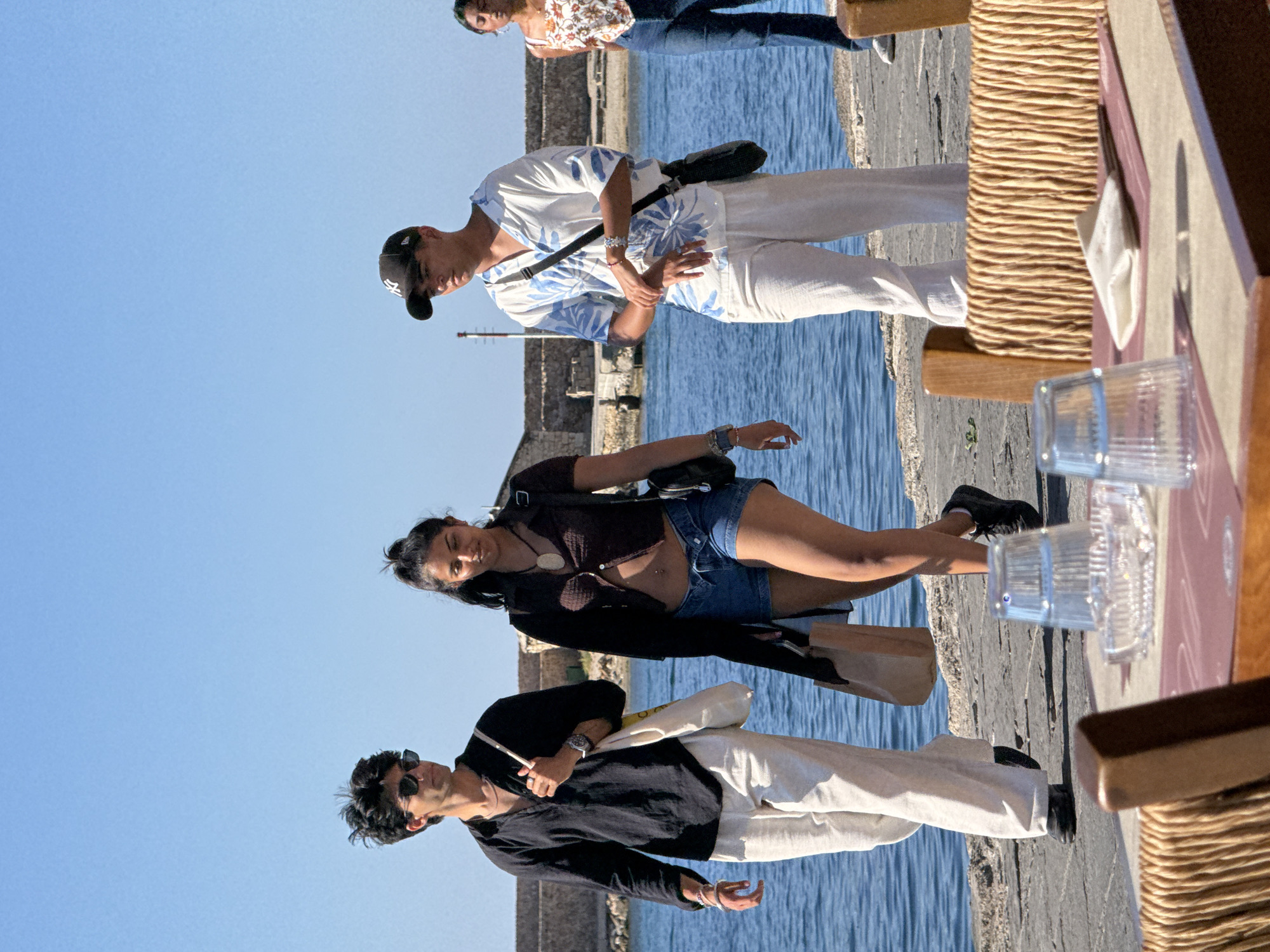 A group of four young adults are walking along a stone waterfront in Chania, Greece. The sky is clear blue and the sea is a deep blue. In the foreground, a table is set with placemats, napkins, and glasses.
The two individuals in the center are a man and a woman walking side-by-side. The man on the left is wearing a black collared shirt, white pants, and sunglasses. He carries a large tote bag over his shoulder. The woman next to him is wearing a dark brown cropped top, denim shorts, and black sneakers. She has a dark shoulder bag and is holding a brown paper bag.
To the right of the central pair, a man is walking with a black baseball cap, a white short-sleeved shirt with a blue floral print, and white pants. He also has a dark shoulder bag. Further to the right, a woman in a floral tank top and blue jeans walks behind.