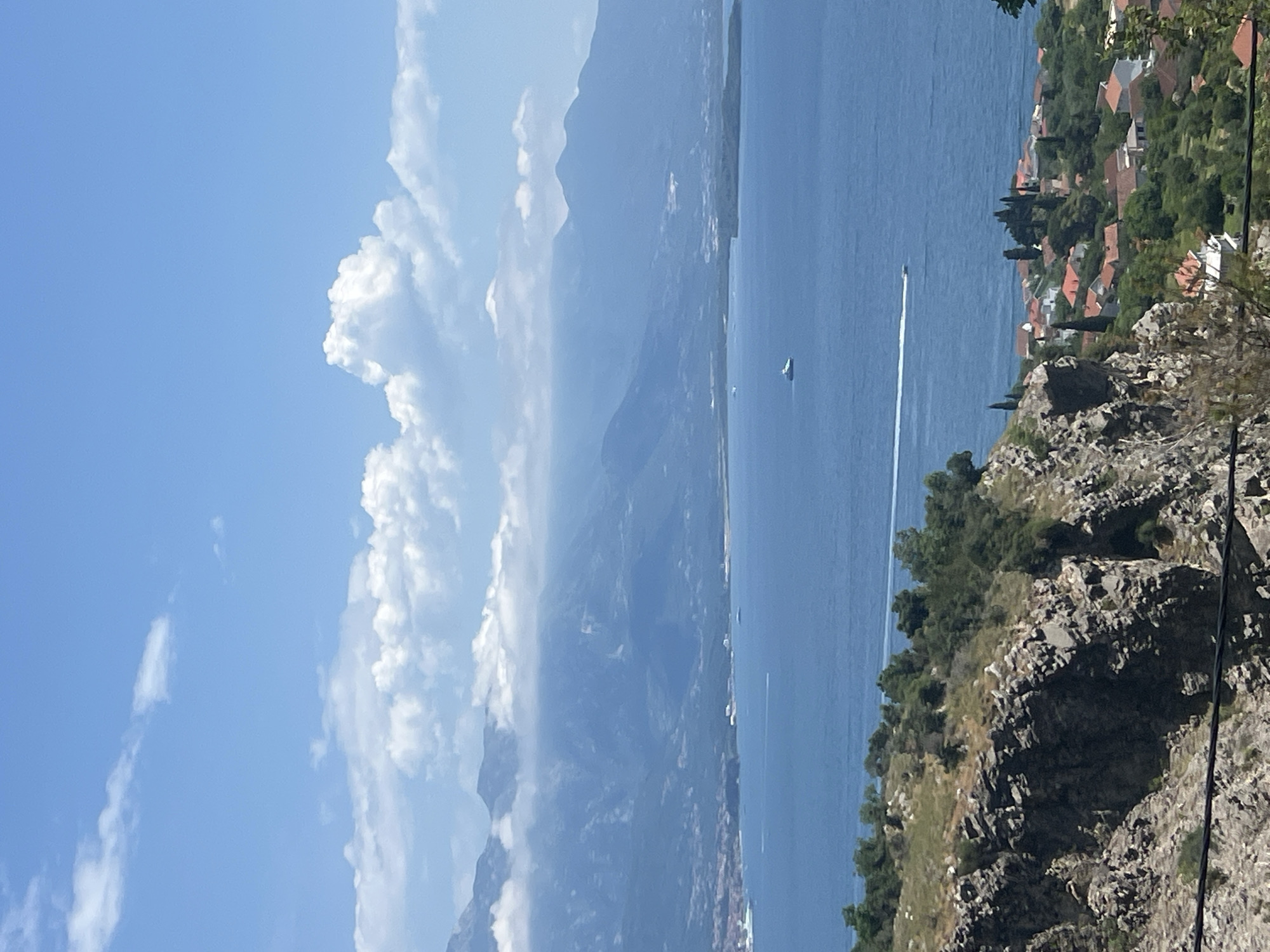 The image depicts a coastal view in Baošići, Montenegro. The Adriatic Sea stretches from the foreground to the horizon, with a boat leaving a white wake. On the right, a rocky hillside is covered with vegetation and interspersed with buildings and red-tiled roofs, indicating residential areas. In the background, mountains rise into a blue sky dotted with white clouds. Utility cables are visible in the lower right corner.