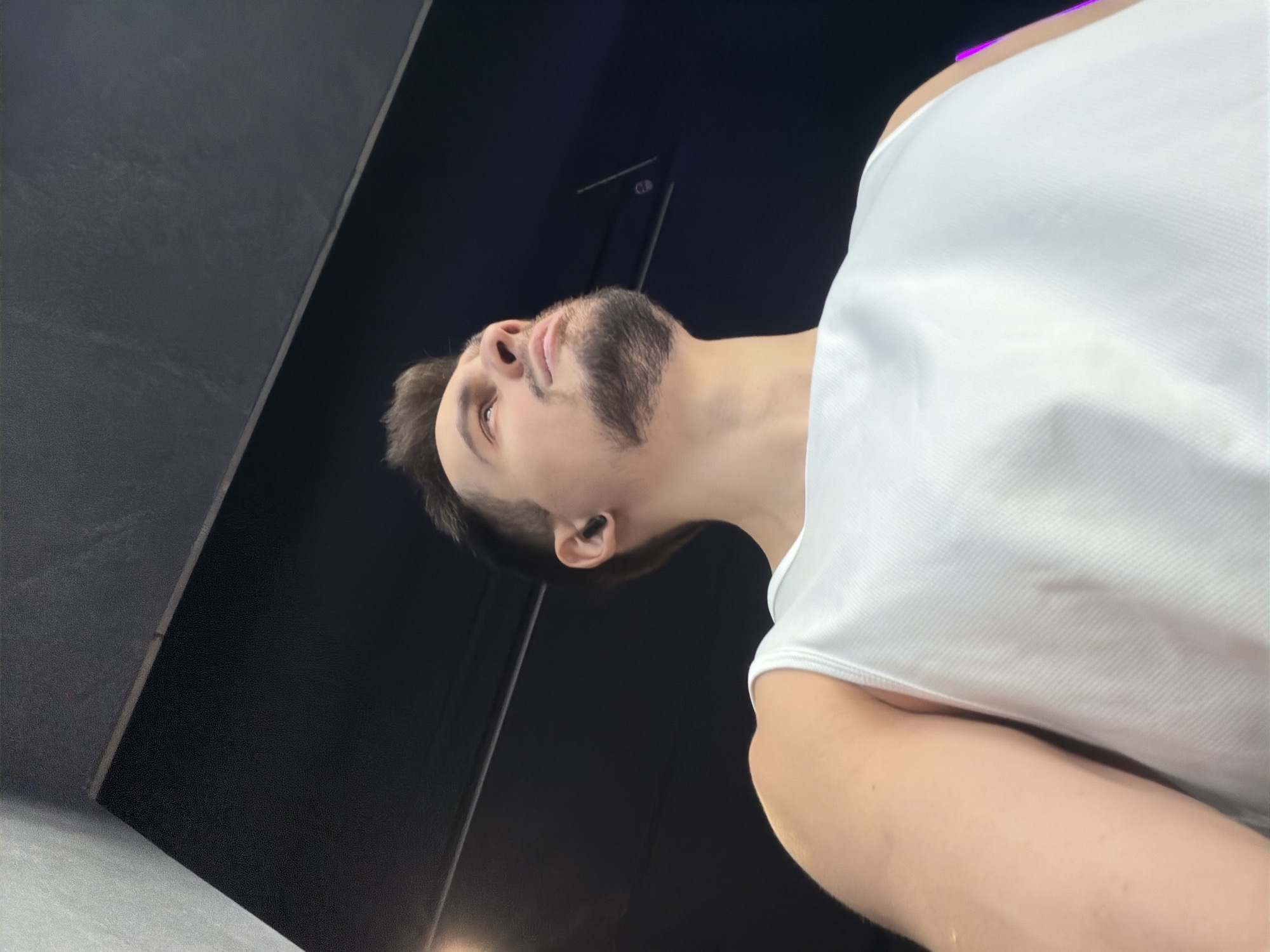 A young man is depicted in a low-angle, close-up shot, gazing upwards and slightly to his left within an indoor setting in Krasnoyarsk, Russia. He appears to be in his 20s or early 30s, sporting short dark hair and a dark, neatly trimmed beard. He wears a white, ribbed sleeveless tank top, which stands out against the predominantly dark background. A black earbud is visible in his left ear. The man's expression is neutral, possibly contemplative or focused, as he looks towards something out of frame above him. The background features sleek, dark surfaces, including a matte black wall and another textured dark grey or black panel, suggesting a modern interior space, potentially a gym or studio. The lighting is artificial, illuminating the man from above. No visible text is present in the image.
