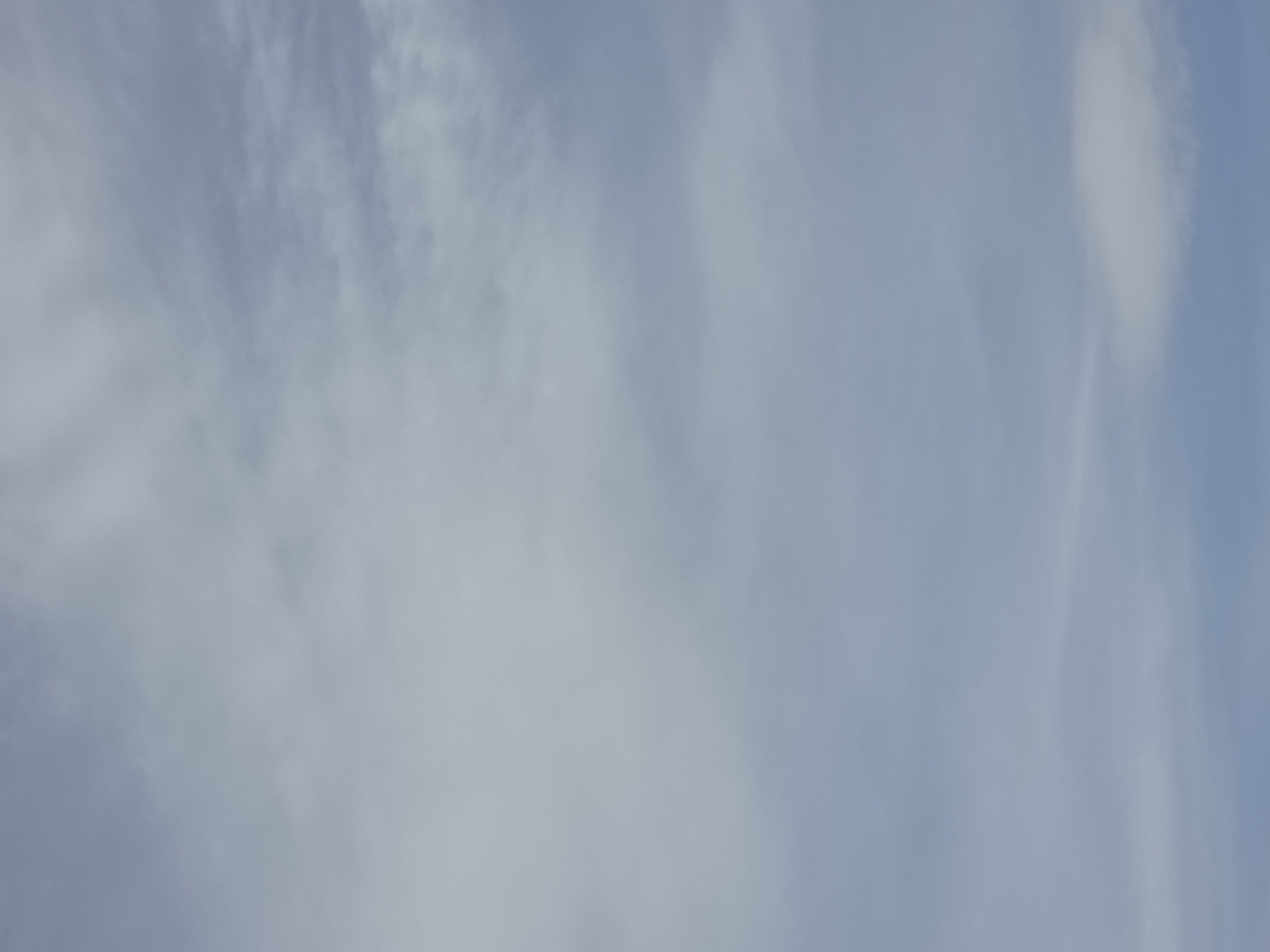The image displays a cloudy sky with a predominantly blue-grey hue. Wispy clouds are scattered across the frame, with some appearing denser than others. The lighting suggests it is daytime. No people, subjects, objects, actions, or interactions are visible. The image appears to be a photograph of the sky. There is no indication of the location being the city of Magnitka, Russia.