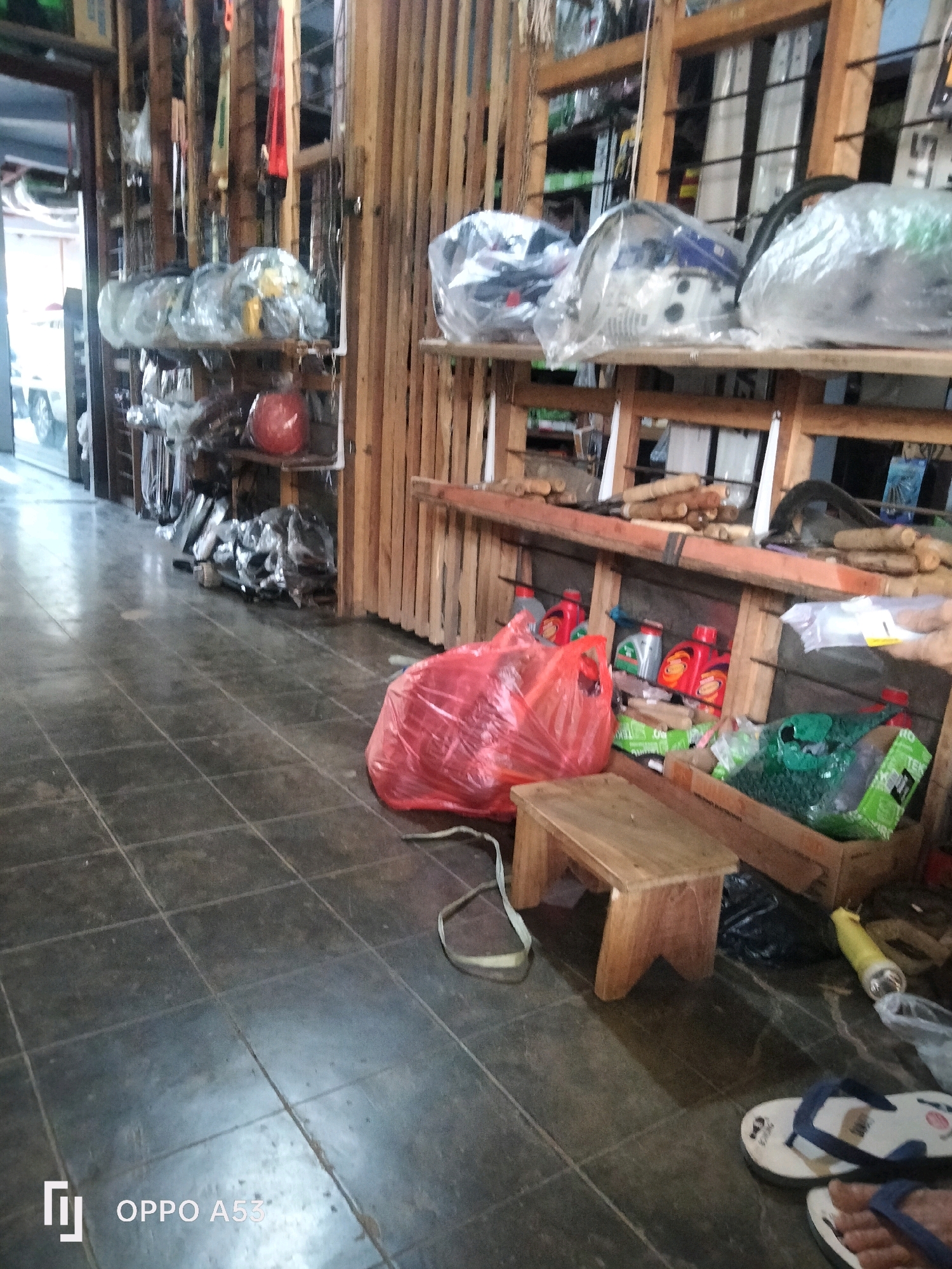 The image depicts an indoor scene, likely a shop, with a tiled floor and wooden shelving units. Various items are stored on the shelves and on the floor. On the shelves, there are objects covered in plastic wrap, some red plastic bottles, and what appear to be engine parts or tools. On the floor, there is a large red plastic bag, a small wooden stool, a brown cardboard box containing green plastic items, a yellow flashlight, and a pair of flip-flops. The lighting is dim, and the overall atmosphere suggests a storage or retail area for mechanical or automotive parts. The presence of the "OPPO A53" watermark in the bottom left corner indicates the image was taken with a mobile phone. This is not Ngulanwetan, Indonesia.