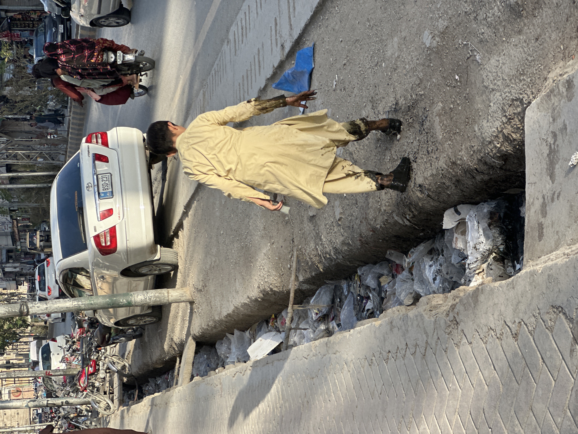 The image depicts a man in a traditional shalwar kameez, partially immersed in a roadside drainage ditch filled with garbage in Quetta City Tehsil, Pakistan. His clothing and boots are visibly soiled with debris and dark stains. He appears to be reaching into the drain with his right hand. A blue object lies on the roadside near him. Adjacent to the drain is a street with parked cars, motorcycles, and pedestrians. The image suggests urban pollution and potential sanitation issues.
