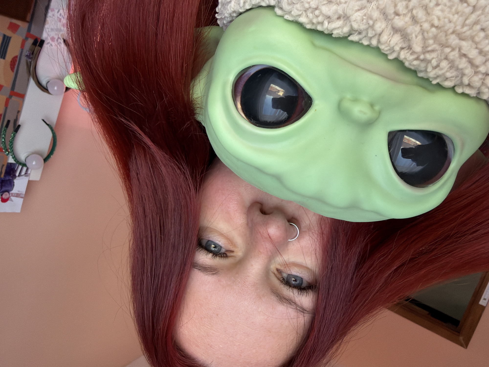 A person with red hair, a nose ring, and blue eyes is holding a "Baby Yoda" toy close to their face. The person and toy are upside down. The toy has large black eyes and a light green face. The toy also appears to have a beige fuzzy hat. The background is a light peach color with items such as a lamp, a postcard, and hair accessories on a surface. A window is visible in the background as well. There is no evidence in the image to determine if this image was taken in Chaikovsky, Russia.
