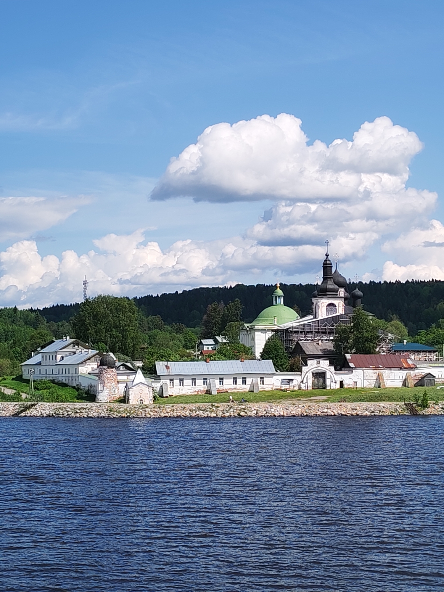 The image shows a tranquil scene of the Goritsky Monastery on the banks of the Volga River near Goritsy, Russia. In the foreground is the rippled surface of the dark blue river. A low rocky shoreline separates the water from the monastery buildings. The monastery is a collection of primarily white buildings with various architectural styles, including green and black domes topped with Orthodox crosses. Scaffolding is visible on one of the church structures, indicating ongoing restoration work. The monastery buildings are framed by a backdrop of lush green trees, providing a contrast to the white structures. A transmission tower is also visible amidst the trees. Above the monastery and trees, a blue sky is punctuated by large, fluffy white clouds.