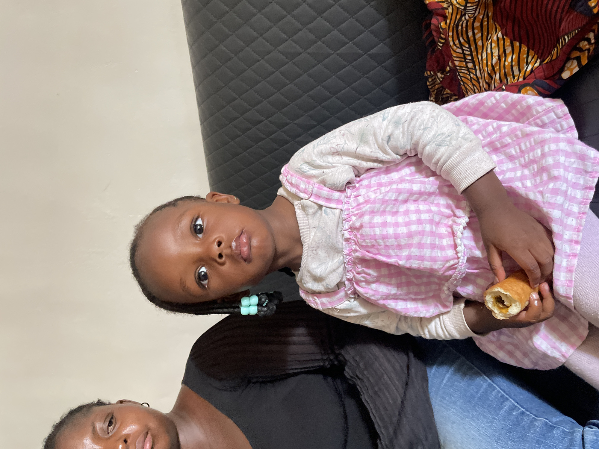 This portrait-oriented image, captured in Barakin Akawo, Nigeria, features a young Black girl and an adult woman, likely a mother figure, indoors. The image is rotated clockwise, but when viewed upright, the child is in the upper part of the frame, looking directly at the camera with a neutral expression. She has dark braided hair, with light blue beads visible on one braid near her ear. She is wearing a light-colored, long-sleeved top underneath a pink and white checkered pinafore dress. The child holds a golden-brown, cylindrical pastry or fried roll in her hands, showing a savory-looking filling. Some residue is visible around her mouth, suggesting she was in the process of eating.
The adult woman is partially visible in the lower part of the frame, beneath the child, wearing a black top and blue jeans. Her face is only partially shown, and she also appears to be looking towards the camera. Both individuals have dark skin tones.
The setting appears to be a domestic interior. Behind the child and adult is a dark-colored sofa or chair with a distinct diamond-quilted pattern. To the upper right, a colorful fabric with traditional African patterns (red, orange, black, and white) is draped over the furniture. The wall in the background is plain and light-colored. The lighting suggests it is daytime. There is no visible text in the image.