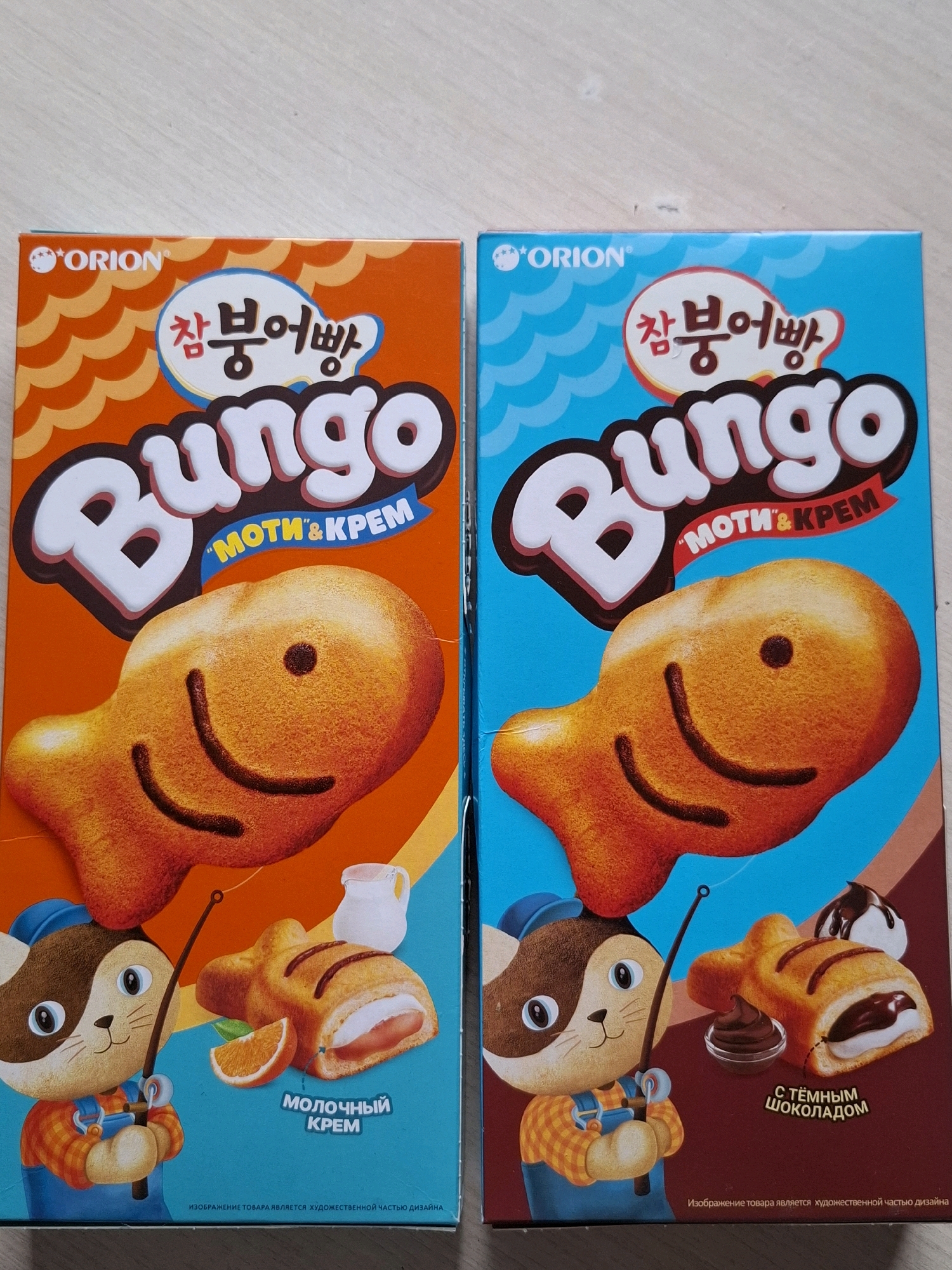 Two boxes of Orion "Bungo" fish-shaped snacks are displayed side-by-side on a light-colored wooden surface, possibly a floor or table. The box on the left is orange and features a drawing of a smiling fish snack filled with cream, alongside a cartoon cat character holding a fishing rod. The text on this box is in Korean and Russian. The box on the right is blue and features a similar smiling fish snack, but this one is filled with chocolate cream, and a similar cartoon cat character. The text on this box is also in Korean and Russian. Both boxes have the "ORION" logo at the top. The image is a product photograph, likely taken indoors. There is no indication of the location being specifically in the city of Tyumen, Russia.