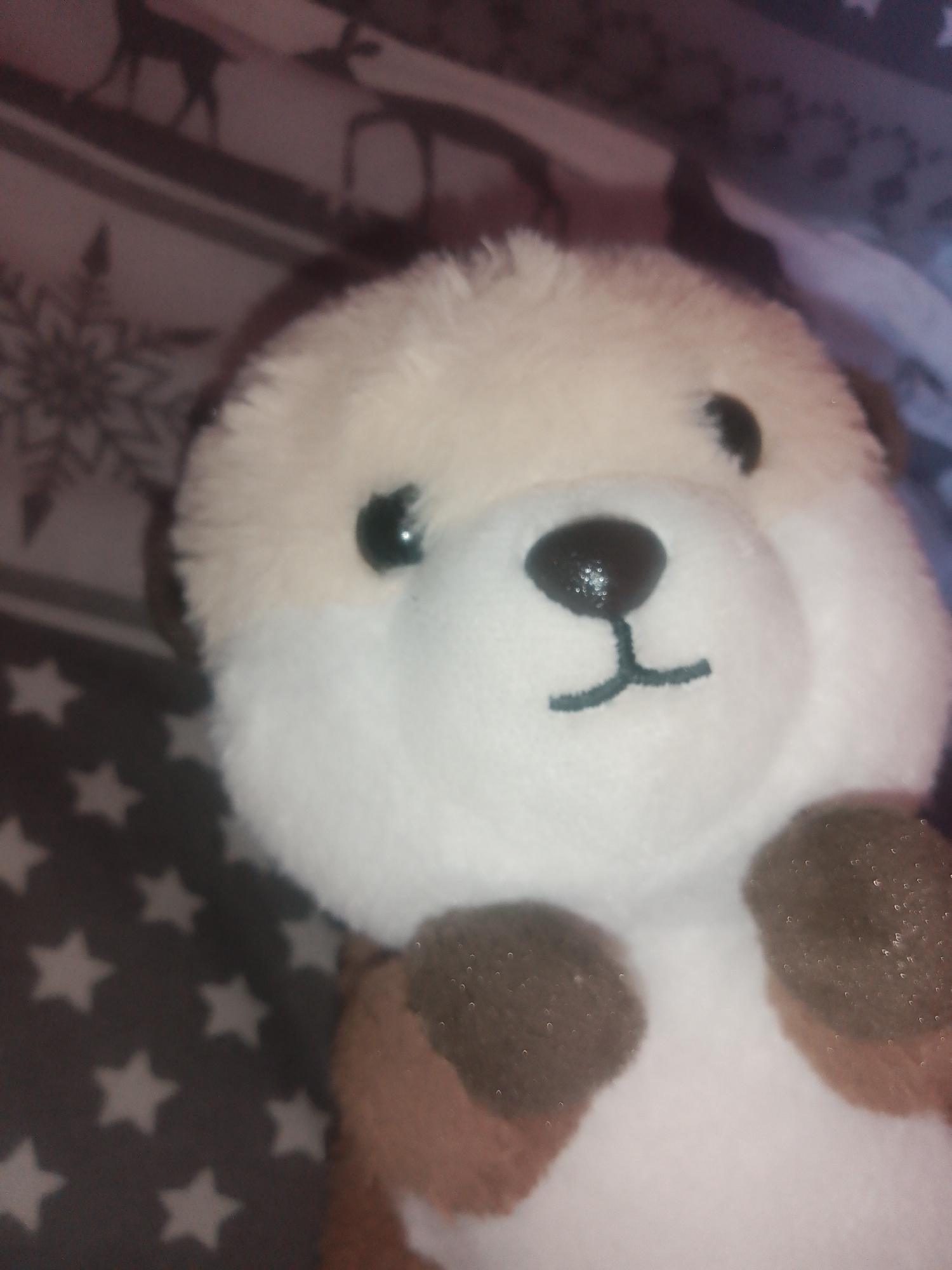 A close-up, slightly blurry image shows a plush toy otter sitting on a patterned fabric. The otter is predominantly white and tan with dark eyes and a dark embroidered nose and mouth. The fabric background has a pattern of white stars on a dark grey base, with some areas showing a blurred pattern of deer and snowflakes. The image appears to be taken indoors, possibly in a domestic setting. There is no indication of the city of Chernivtsi, Ukraine, or any specific location.