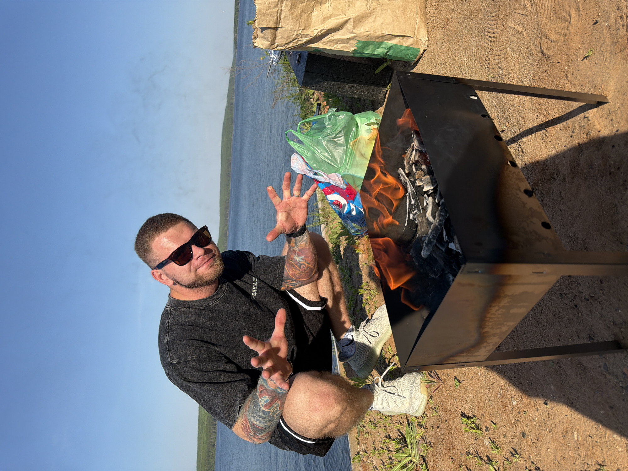 A man with a beard, sunglasses, and tattoos on his arms is sitting cross-legged next to a metal grill with flames in it. He is facing forward, holding his hands up in a gesture. Next to the grill is a brown paper bag and a green plastic bag with what appears to be clothing inside. The location appears to be near the water, possibly the reservoir next to the city of Chaikovsky, Russia. The ground is dirt and there is some sparse vegetation. The sky is mostly clear.