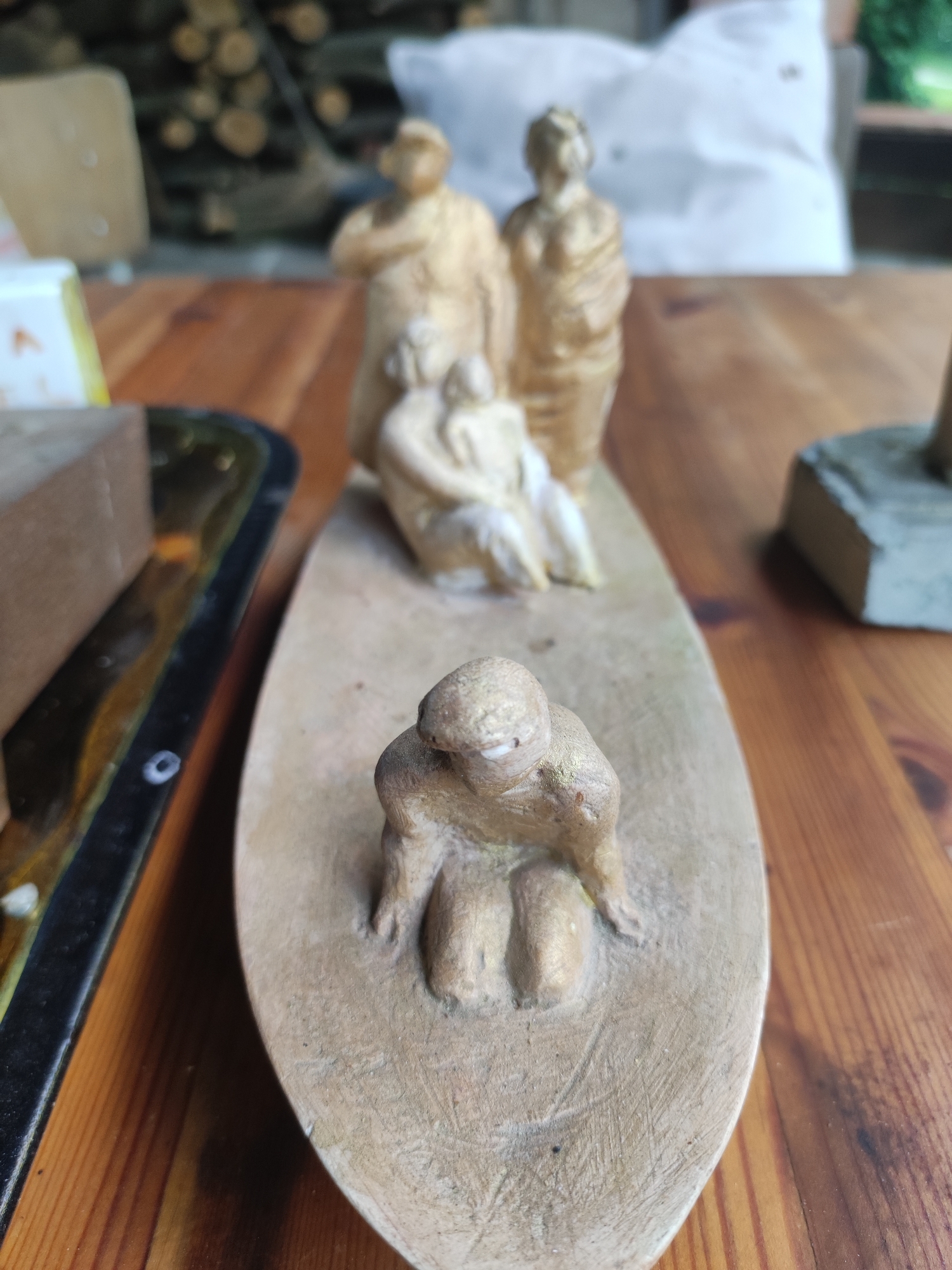 The image shows a collection of small, beige-colored sculptures of human figures, likely made of wood or clay, arranged on a wooden table. In the foreground, a single figure is kneeling on an oval base. Behind this figure, a group of three figures is depicted, two standing and one seated with a child. The background is blurred, showing stacked firewood and a white cloth. To the left of the sculptures is a black tray with a dark object, possibly a block of wood. To the right, a gray rectangular object is visible. The overall setting appears to be outdoors or in a workshop, given the presence of natural materials. There is no indication of a specific location within the city of Dresden, Germany.