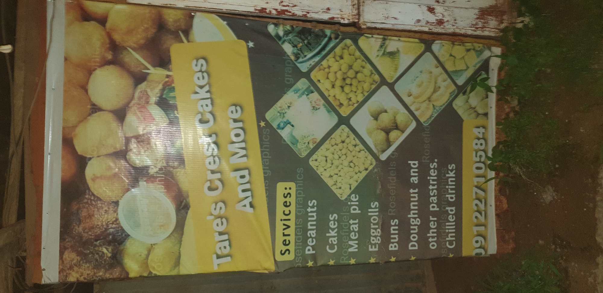 A weathered banner advertising "Tare's Crest Cakes And More" is mounted on a rough, wooden structure, likely an exterior wall or fence, in Narayi, Nigeria. The banner, predominantly yellow and dark gray, features images of baked goods and a list of services. The services listed include "Peanuts," "Cakes," "Meat pie," "Eggrolls," "Buns," "Doughnut and other pastries," and "Chilled drinks." A phone number, "09122710584," is prominently displayed at the bottom. The banner itself is creased and shows signs of age and exposure to the elements. The background is out of focus but suggests a dimly lit, outdoor environment with some foliage and possibly more wooden structures. No people are visible in the image.