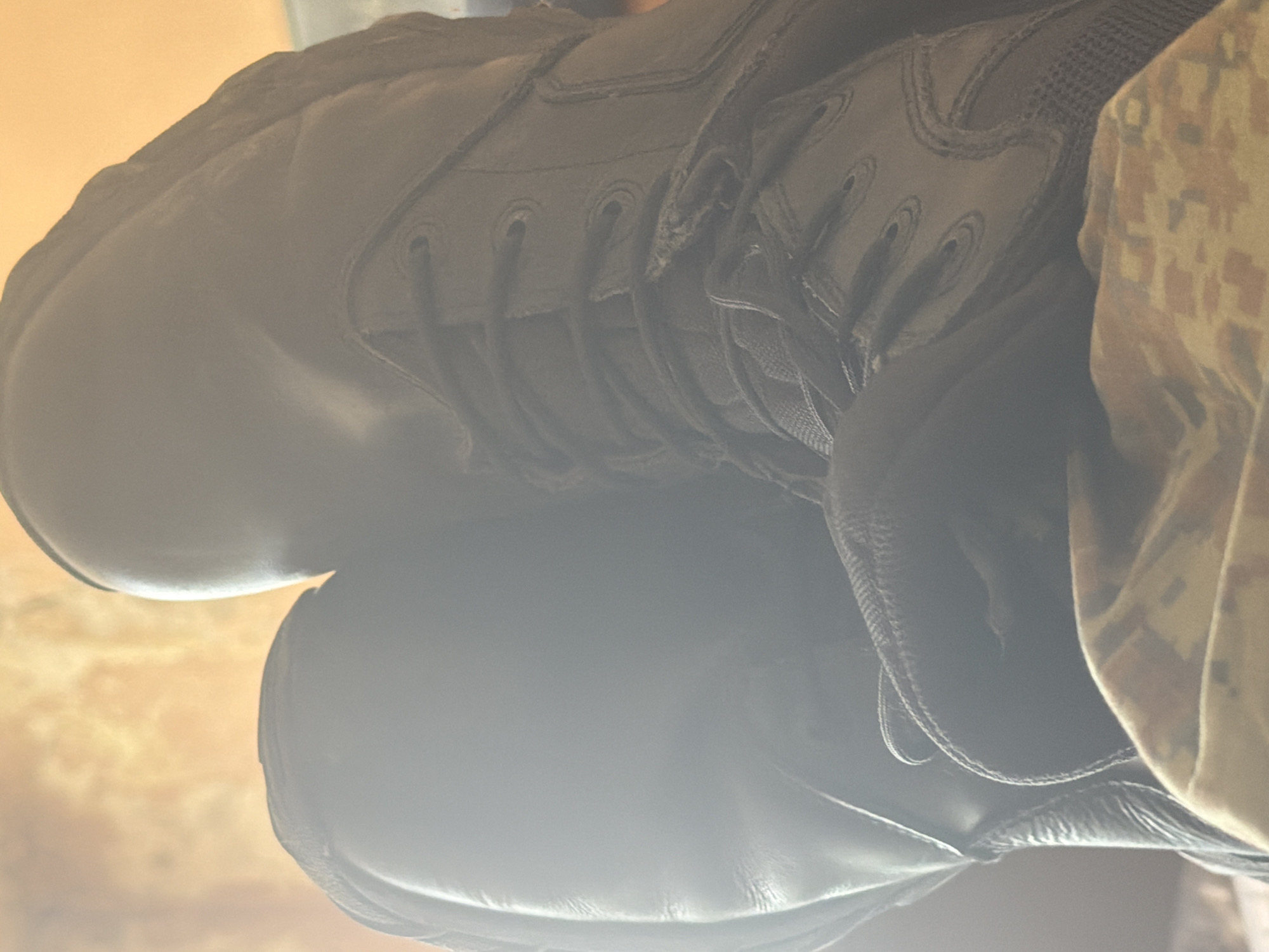 The image shows a close-up of two black leather boots, with one boot placed on top of the other. A camouflage-patterned garment is visible to the right of the boots. The laces of the top boot are visible. The image is taken with low lighting and unclear focus. There is no information that indicates this image was taken in Dolna Mitropolia, Bulgaria.
