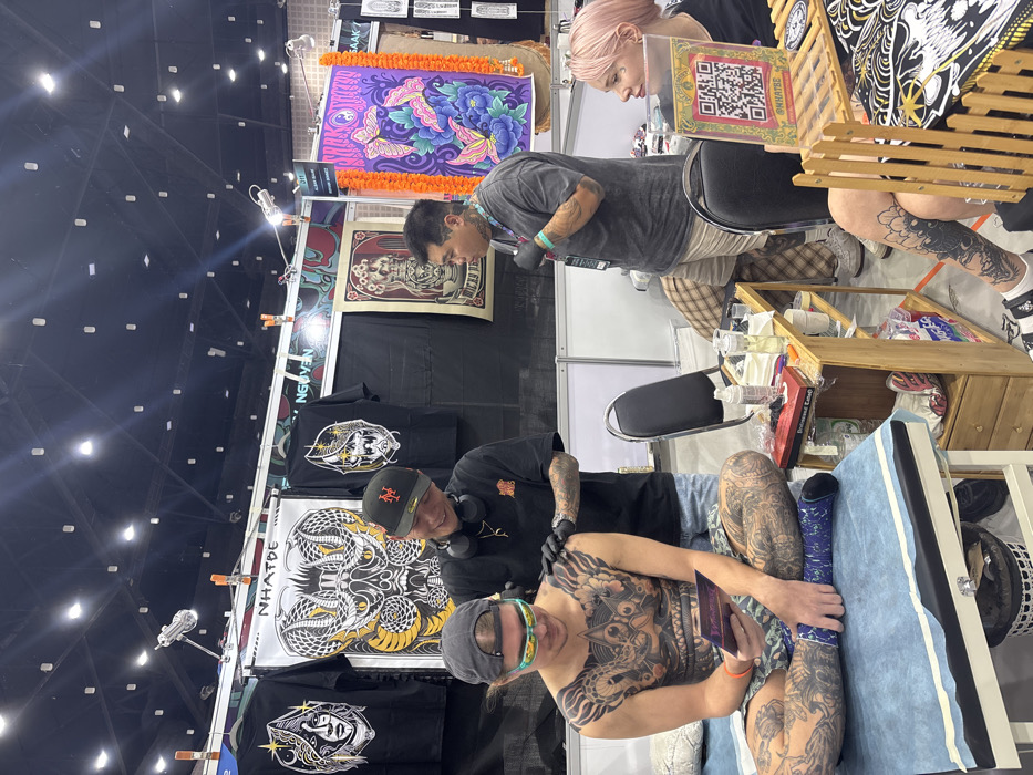 The scene is an indoor tattoo booth at a convention in the city of Ban Rong Ruea Kham, Thailand. Four individuals are present. In the foreground-left, a male client, heavily tattooed across his torso and legs, sits cross-legged on a light blue examination table, wearing a grey cap, reflective green sunglasses, and camouflage shorts with a blue compression wrap on his left leg. He holds a small, dark object and looks down, smiling slightly. Behind him and to his right, a male tattoo artist, wearing a black cap, black t-shirt, denim shorts, and black gloves, applies a tattoo to the client's right shoulder with a machine, smiling at the client. To the right, a second male, also tattooed, stands wearing a grey t-shirt, plaid shorts, and black gloves, with an ID badge, looking rightward. Further right, a female with pink hair and a small facial tattoo is seated at a table, observing. A clear acrylic stand displaying a QR code for "@@NHATBE" is positioned on the table in front of her. A two-shelf wooden utility cart holds tattoo supplies, including bottles, liquids, and a red box, situated between the client and the seated female. The booth's rear wall displays black t-shirts featuring designs such as a stylized face, intertwined cobras with "NHATDE" text, and a skull. Also visible are a poster with floral vase motifs and a vibrant purple banner with butterfly designs. The overhead ceiling features exposed structure and numerous track lighting fixtures.