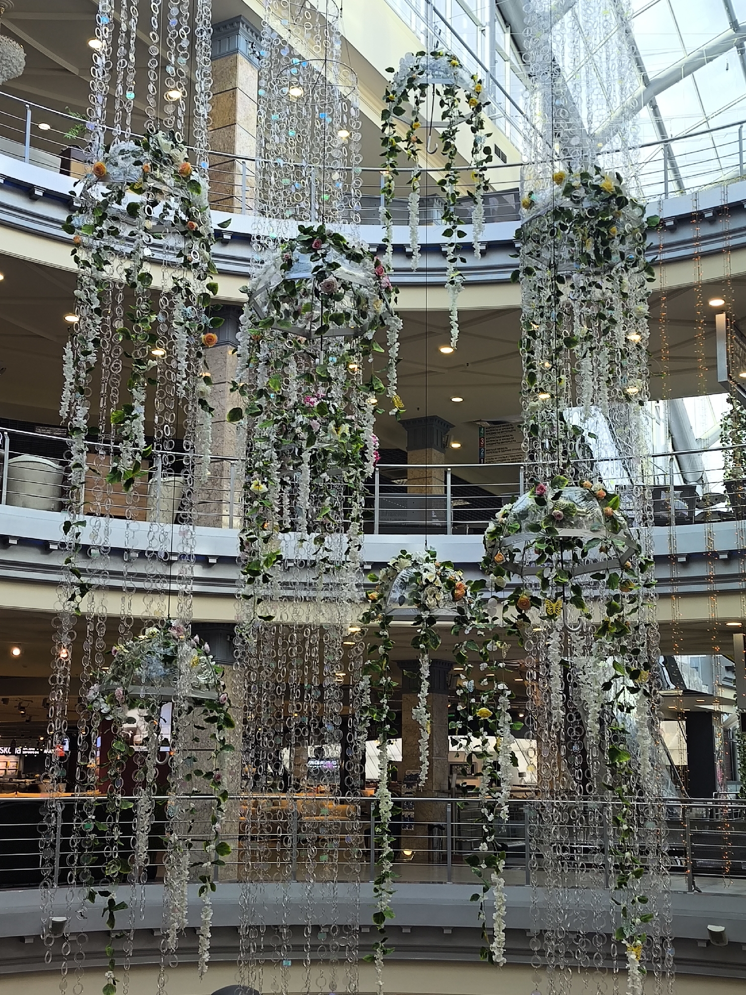 This image depicts the interior of a large, multi-level building, likely a shopping mall or an atrium, located in Khimki, Russia. The central space is adorned with an elaborate hanging decorative installation. Numerous vertical strands of clear, reflective discs or beads cascade from the ceiling, creating a shimmering curtain effect.
Interspersed within these strands are several large, floral arrangements that feature green foliage, white flowers, and colorful blooms such as pink and orange roses, and yellow flowers. Many of these floral elements appear to be draped over what look like inverted clear umbrellas or dome-shaped structures. Additional white cascading flowers and small decorative butterflies are also visible within the foliage.
The background reveals the building's contemporary architecture, including multiple floors connected by metal railings. A prominent glass skylight or ceiling allows natural light to illuminate the space, complemented by various recessed artificial lights. Reflective surfaces, possibly mirrored walls or glass, create blurred reflections of the decorations.
No people are visible in the scene. On a wall in the background, a sign with Russian text is partially discernible, stating "ОХРАНЯЕТСЯ ТЕРРИТОРИЯ" (TERRITORY IS GUARDED) and "ПОД ВИДЕОНАБЛЮДЕНИЕМ" (UNDER VIDEO SURVEILLANCE), along with "ВИДЕОКАМЕРАМИ ВЕДЕТСЯ" (VIDEO CAMERAS ARE CONDUCTING) and "ВИДЕОСЪЕМКА" (VIDEO SHOOTING). Other visible text fragments include "SKLA" and "AMATOP," likely part of store names. The decorations suggest a festive or elegant ambiance, possibly for a seasonal event or as a permanent artistic display.
