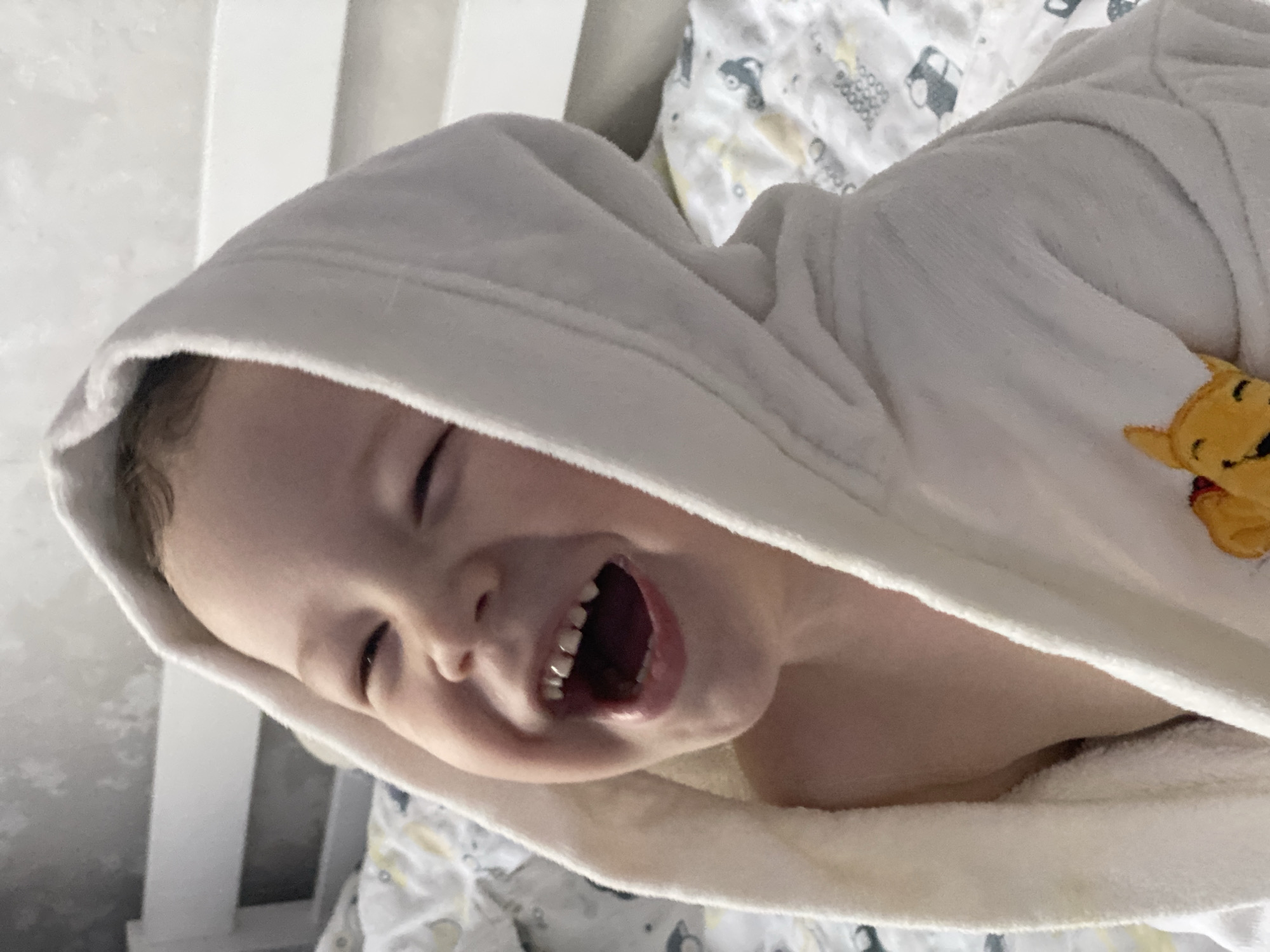 An original, slightly blurry, and rotated portrait-style photograph captures a joyful young child, likely a toddler or preschooler, in an indoor setting. The child, with light skin and head positioned to the left, is seen from a slightly elevated perspective, laughing heartily with their mouth wide open and eyes closed in amusement, showcasing several small teeth. They are wearing a white, hooded bathrobe, possibly made of terry cloth, which features a yellow Winnie the Pooh appliqué on the right side of the chest area. Part of a light-colored, patterned sheet or blanket with cartoon cars and geometric shapes is visible in the background and beneath the child, suggesting they are in a bed or crib, possibly indicated by a white railing-like structure on the far left. The scene evokes a strong sense of playful happiness and genuine, uninhibited joy, possibly captured after a bath or during a lively moment of play. The photograph was taken in Sochi, Russia. No visible text is present in the image.