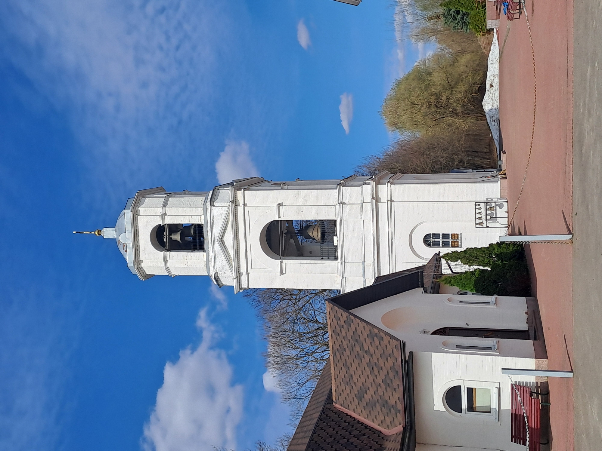 The image captures a white Orthodox church bell tower against a blue sky with scattered clouds. The tower has multiple tiers with arched openings revealing large bells. A brown tiled roof of a smaller building is visible at the base of the tower, and part of a white building adjoins it. Trees are visible in the background. A red brick-paved area and a white wall run along the right edge of the image. The location is identified as Ivashevo, Russia.