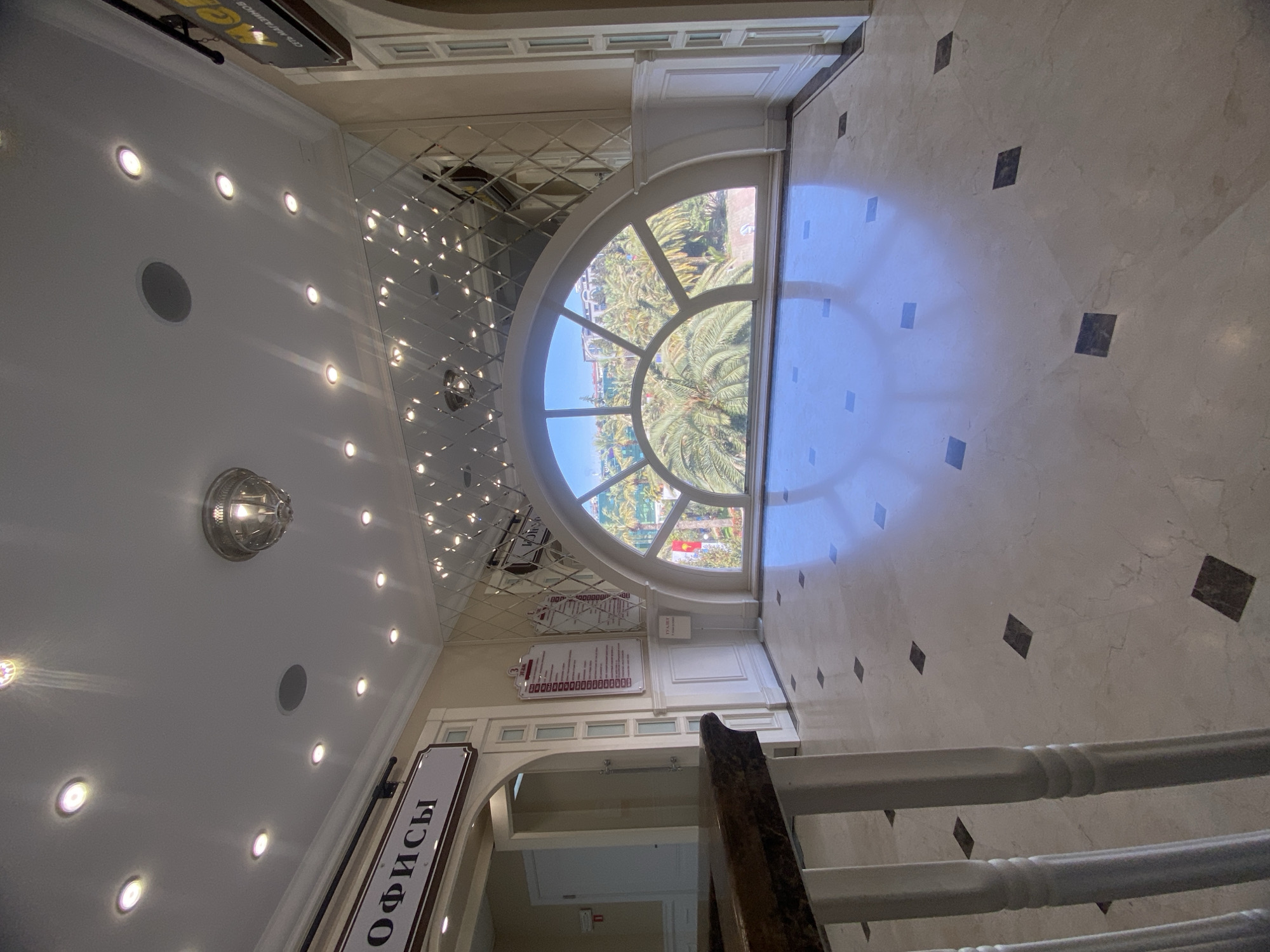 The image depicts an interior space, likely in a building in Sochi, Russia, as indicated by the Cyrillic inscription "ОФИСЫ" (Offices) on a sign. The view is from a slightly elevated position, perhaps a landing on a staircase, with white railings visible in the foreground.
The interior features a glossy, marble-like floor with dark square inlays. A large, arched window dominates the scene, offering a view of a tropical landscape with palm trees and buildings. The reflection of the window in the floor creates a bright, almost blinding effect.
The ceiling is white and illuminated by numerous recessed lights, casting a soft glow. The upper part of the wall adjacent to the window is covered in a diamond-patterned mirror, reflecting the lights and contributing to the bright atmosphere. There are also some framed notices with text on the wall near the window.