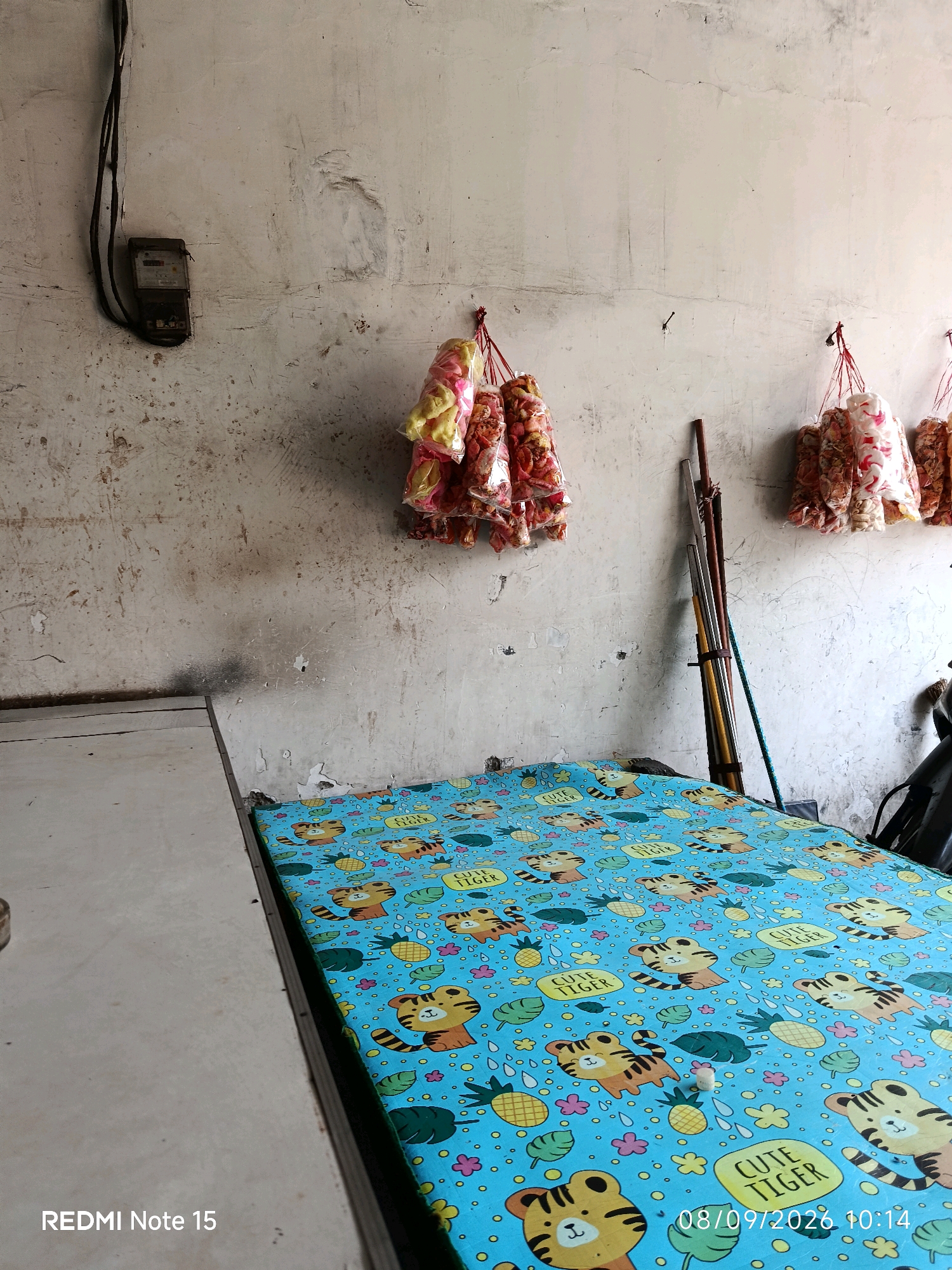 The image captures an indoor scene, likely a simple room or a shop, in Keboansikep, Indonesia. The foreground features a mattress with a vibrant blue cover adorned with cartoon tigers and the text "CUTE TIGER." To the left, a portion of a light-colored surface, possibly a table or counter, is visible. The background reveals a textured, off-white wall that shows signs of age and wear, with a visible electrical meter box mounted on the upper left. Several bunches of packaged snacks, in red and yellow hues, are hanging from the wall, suspended by red strings. Leaning against the wall in the mid-ground are several long poles or rods, possibly tools or broom handles, along with a blue rope. The overall impression is of a functional, unadorned space that might serve a commercial purpose, possibly a small convenience store or a residence with a business operating within it.