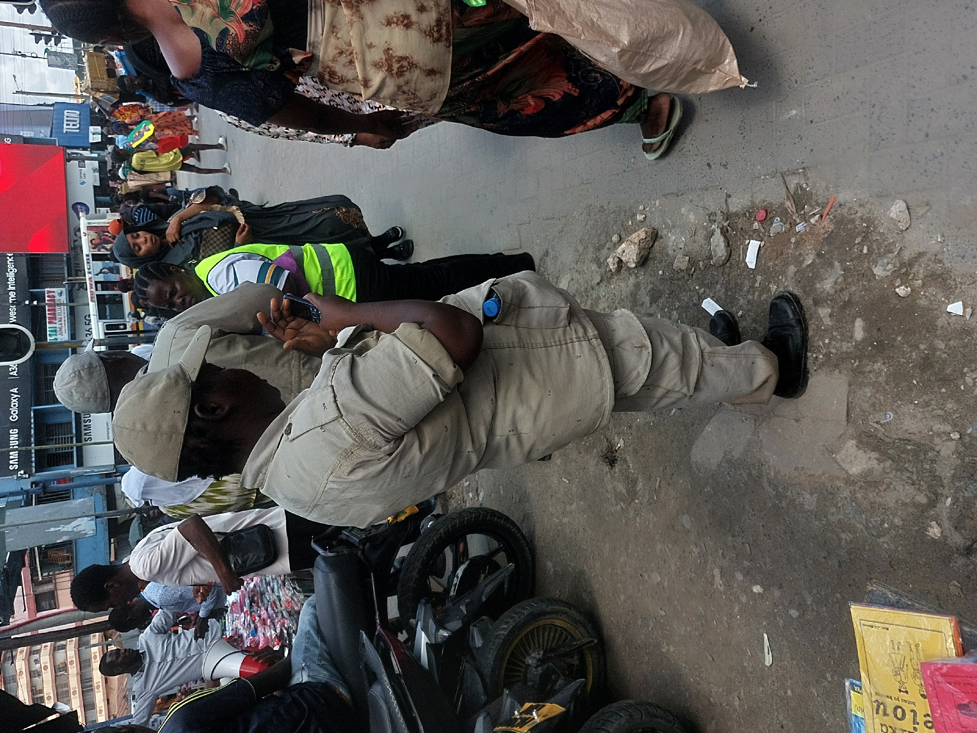 This image captures a bustling urban street scene, characteristic of Dar es-Salaam, Tanzania, during daytime. The perspective is slightly elevated, looking down at a crowded thoroughfare filled with people, vehicles, and commercial signage.
In the foreground, a man in a light beige uniform and matching cap, likely a security guard or public service worker, is depicted from the waist up, holding a dark smartphone. His uniform features repeating "RFC" and "KJA" patterns, and a blue cap is tucked into his left chest pocket. Adjacent to him, another man in a similar uniform is partially visible.
The street teems with numerous individuals engaged in daily activities. A woman wearing a dark head covering and dress stands in the mid-ground, near another person in a bright neon green safety vest. Further down the street, people are walking and standing, and one individual can be seen holding a megaphone, suggesting a public announcement or event.
The environment is distinctly commercial, with several motorcycles parked or navigating the street. The ground comprises a mix of concrete and unpaved dirt, scattered with small debris. Prominent advertisements and signs line the left side of the frame, displaying text including "SAMSUNG Galaxy A A30", "west me Intelligence", "JUMLA NA REJAJA" (Swahili for wholesale and retail), "TECTO", "36.5G", "BOWAICHI", and "STAI PHARMACY". In the bottom right, a stack of items, possibly books, includes a yellow one with a drawing of people and the words "hotuo yo kwa" and "PYA".
The scene conveys the energy of a typical, busy urban setting, with diverse activities unfolding under what appears to be an overcast sky or during late afternoon.