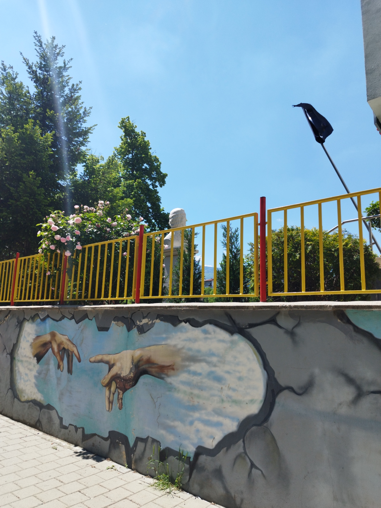 The image captures a street scene in Sofia, Bulgaria, featuring a low concrete wall with a mural depicting the hands from Michelangelo's "The Creation of Adam." Above the wall, a yellow fence with red posts runs along the top. Behind the fence, there is a statue of a bust, possibly of a historical figure. A lush green tree with pink flowers is visible to the left of the statue. A black flag flies high on a pole extending from behind the fence. The sky is clear and blue, with bright sunlight. The ground is paved with interlocking gray stones.