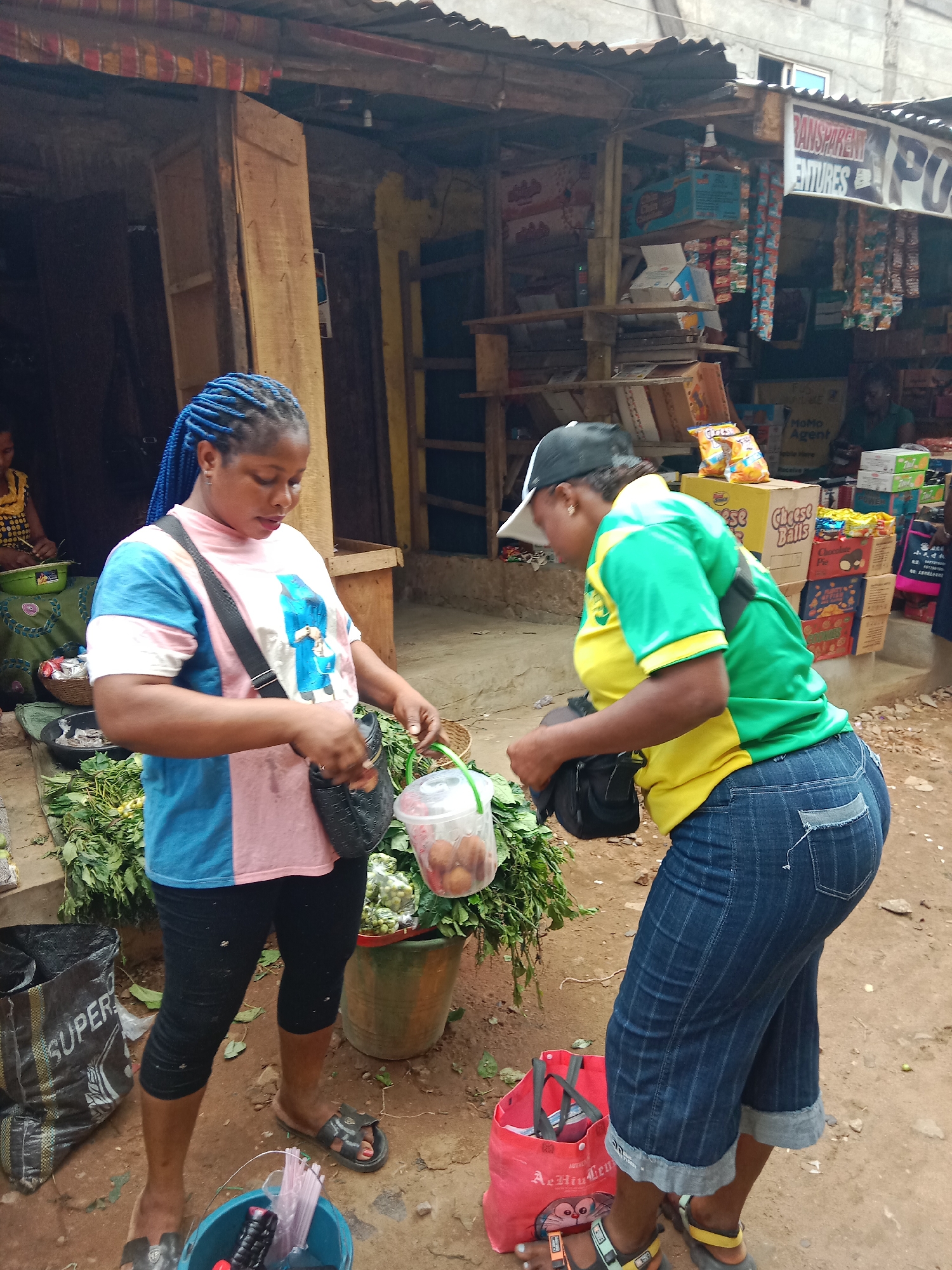This image depicts a bustling outdoor market scene in Onitsha, Nigeria, with two women as the primary subjects. The woman on the left, with vibrant blue braided hair, wears a pink and blue color-block t-shirt, black capri pants, and sandals. She is holding a clear plastic container with fruit, possibly garden eggs, and appears to be retrieving something from her black shoulder bag. Around her feet are large quantities of green leafy vegetables and a black bag labeled "SUPER."
The second woman, slightly turned away from the camera, wears a yellow and green t-shirt, blue denim capri jeans with distinctive stitching and a distressed back pocket, and a black baseball cap. She also wears a black shoulder bag and is focused on the interaction, looking down at her hands or the space between them. A red bag printed with "AiHuiLen" is visible on the ground near her feet. In the background, a third woman is partially visible at a stall to the far left.
The setting is characterized by makeshift market stalls constructed from wood and corrugated metal roofs. Various goods are displayed, including packaged snacks like "Cheese Balls" and "Chocolove Pie," indicating a general store setup. Visible signs in the background read "TRANSPARENT VENTURES" and "MoMo Agent," suggesting business names and services. The ground is reddish-brown dirt, indicating an unpaved outdoor environment. The scene captures a typical daytime commercial activity in an African market.