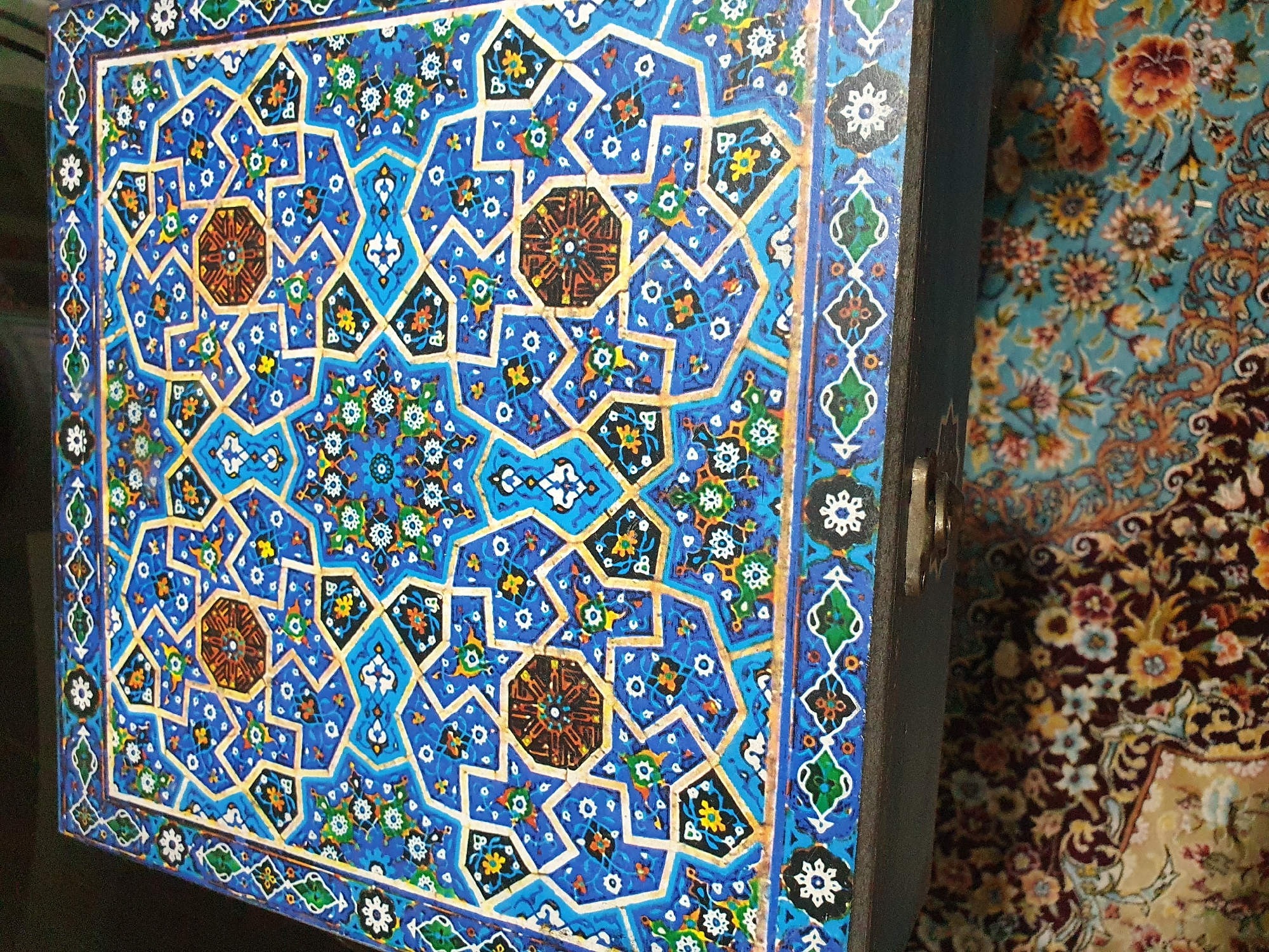 The image shows a close-up of the top surface of a decorative wooden box, adorned with intricate blue and gold geometric patterns and floral motifs. The box is placed on what appears to be a carpet with a floral design. A metal drawer pull is visible on the front edge of the box. The overall impression is one of craftsmanship and cultural artistry. There is no visible indication of the city of Shiraz, Iran, in the image itself.