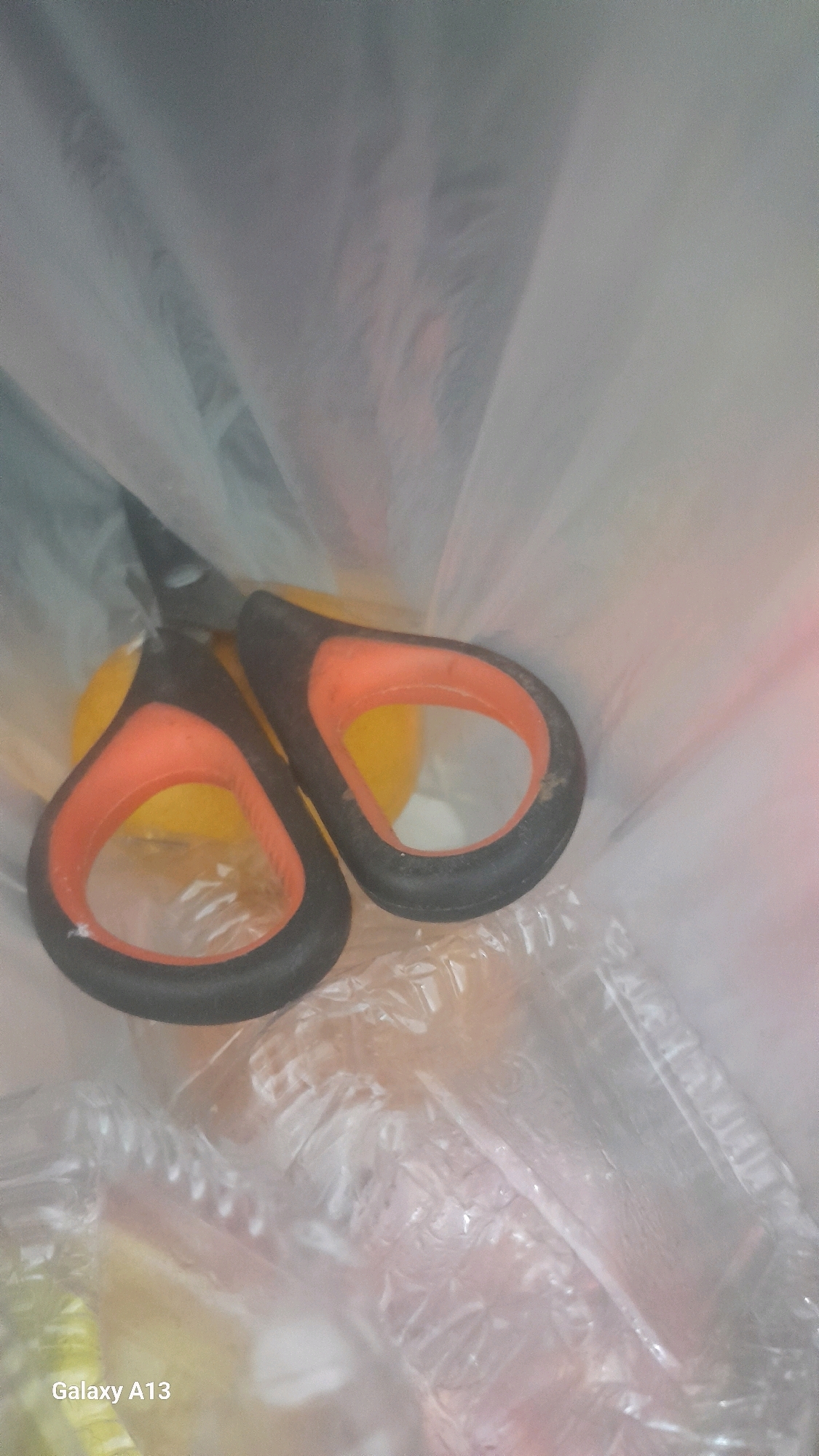A pair of black and orange handled scissors are lying in a clear plastic bag. The scissors are positioned diagonally, with the handles facing the viewer and the blades pointing upwards and to the left. A yellow object, possibly a fruit or vegetable, is partially visible beneath the scissors. The bag appears to be transparent and somewhat crinkled, obscuring some of the details within. The overall lighting is diffused and appears to be artificial. The bottom left corner of the image shows the text "Galaxy A13" which is likely a watermark from the device used to capture the photograph. The scene appears to be indoors.