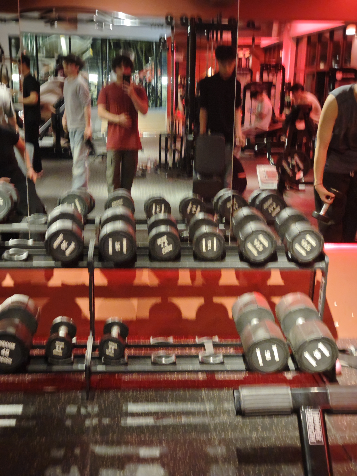 The image depicts the interior of a fitness center located in the Chongqing High-tech Zone, China. The scene is dominated by a reflective wall, likely a mirror, which creates a complex series of reflections. In the immediate foreground, two parallel rows of black dumbbells with silver handles are neatly arranged on a multi-tiered rack. Visible markings on the dumbbells include "10 LB," "15 LB," and "40 LB," among others.
In the midground, seen through the mirror, at least five male individuals are present. One male, positioned centrally, is actively taking a mirror selfie with a smartphone, obscuring his face. He wears a red short-sleeved shirt and dark trousers. To his left in the reflection, another male wears a light grey long-sleeved shirt and light-colored pants, standing with his gaze directed leftward. Further left, a male in a black short-sleeved shirt and dark pants is visible. To the right of the central figure, a male's torso and arm are visible, holding a small dumbbell. He wears a dark top. Additional indistinct figures are present deeper in the reflected background, interacting with various gym equipment.
The environment is characterized by a strong reddish light source on the right side, casting a red glow on the walls and ceiling. Numerous other gym machines, including weight stacks and benches, are visible in the reflections. Large windows in the far background reveal green foliage outside, suggesting the gym is situated with an outdoor view. The floor consists of dark, textured gym matting. The overall impression is an active, well-equipped gym under dim, atmospherically lit conditions.