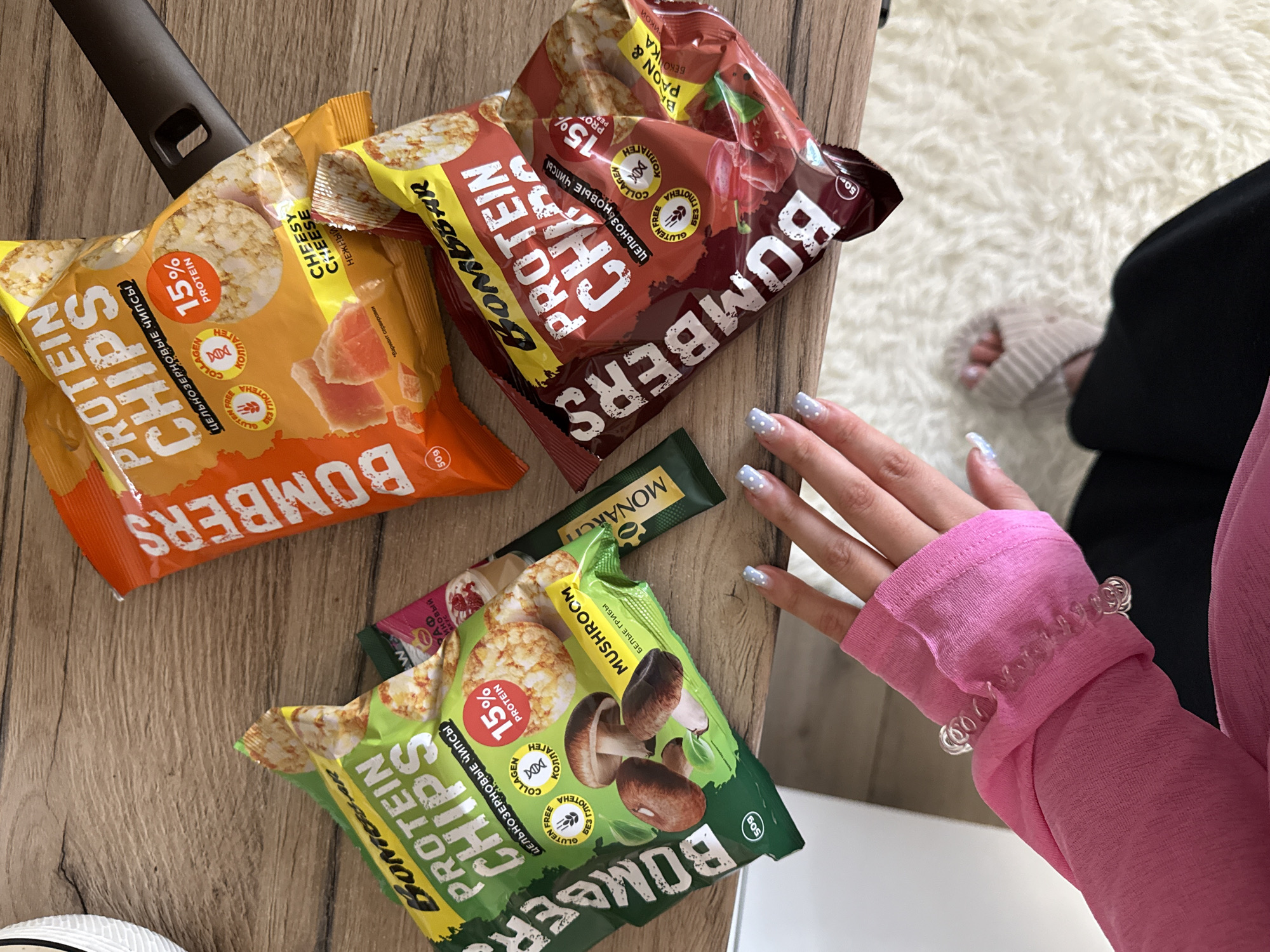 A close-up overhead shot shows several bags of "Bomber Protein Chips" arranged on a wooden surface, possibly a table, next to a fluffy white rug. The bags are primarily orange, red, and green, with prominent text in English and Russian advertising "Protein Chips" and flavors like "Cheesy Cheese," "Becons & Ricoh," and "Mushroom." Some bags indicate "15% Protein," "Gluten Free," and "Collagen."
In the foreground, a person's hand, adorned with light blue nail polish with white polka dots, is reaching towards the bags. The person is wearing a bright pink long-sleeved top with a ruffled cuff, and a clear, coiled bracelet is visible on their wrist. Part of the person's arm and shoulder are in view, along with a glimpse of their legs and feet, wearing beige socks, on the rug. The lighting suggests it's daytime. The scene appears to be indoors, possibly in a living room or kitchen area in Tliustenkhabl', Russia, given the location context. The arrangement of the snack bags and the presence of the hand suggest a casual moment of someone about to enjoy a snack.
