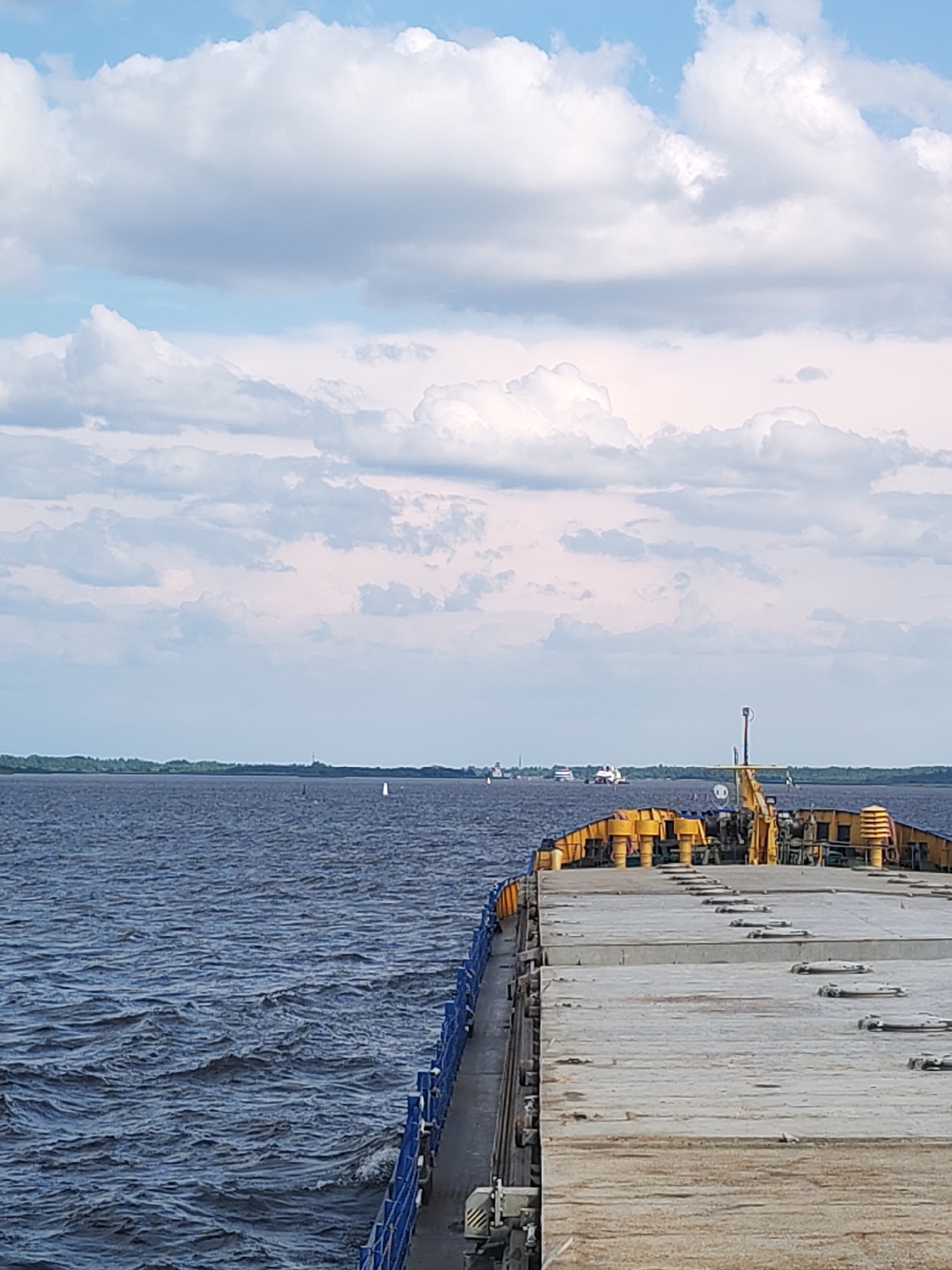 This image captures a view from a vessel on a body of water, likely the Sheksna River near Krokhino, Russia, given the presence of the remnants of the overgrown dam of the former Krokhino Hydroelectric Power Station, which is now a tourist attraction. The foreground is dominated by the deck of the vessel, a flat-topped barge with a concrete surface and various metal fixtures. The barge's railing, painted blue, runs along the left side.
The water is a deep blue-gray, with visible ripples and small waves indicating some movement. In the mid-ground, a distant shoreline is visible, characterized by a strip of green trees. Several other vessels are present on the water, including what appear to be barges or other cargo ships. One vessel in the distance features a distinctive red and white superstructure.
The sky above is filled with a mix of white and gray clouds, with patches of blue sky visible. The lighting suggests it is daytime, with diffused light due to the cloud cover. The overall scene conveys a sense of travel or transport on a large waterway.