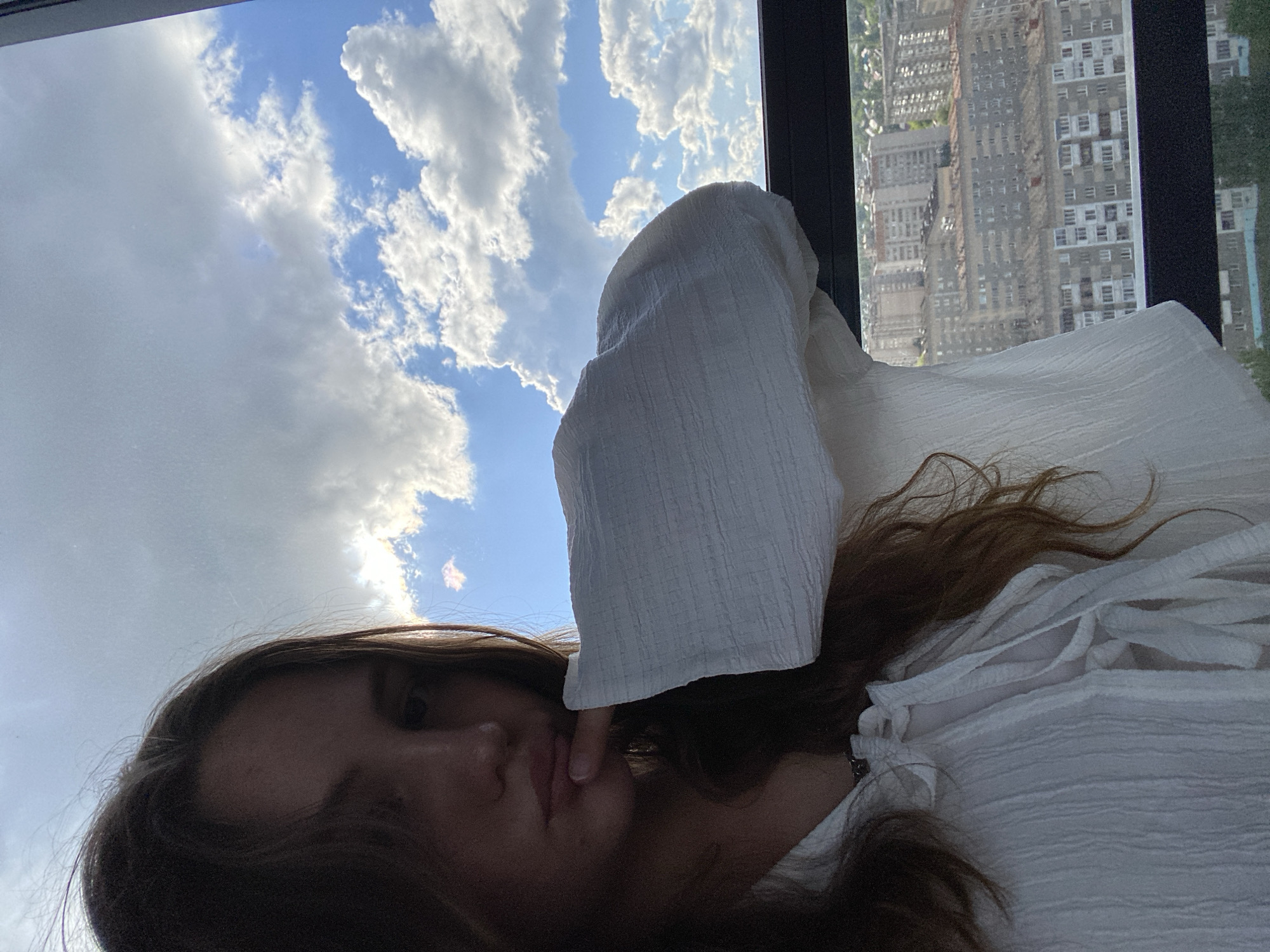 A woman is photographed from a low angle, with a view of the sky and city buildings visible through a window behind her. The woman has light skin, brown hair, and is wearing a white top. Her face is partially obscured, and her finger is touching her lips. A white hooded garment is draped over her head. The sky is blue with puffy white clouds. Outside the window are tall buildings. The location of the image is not in the city of Izhevsk, Russia.