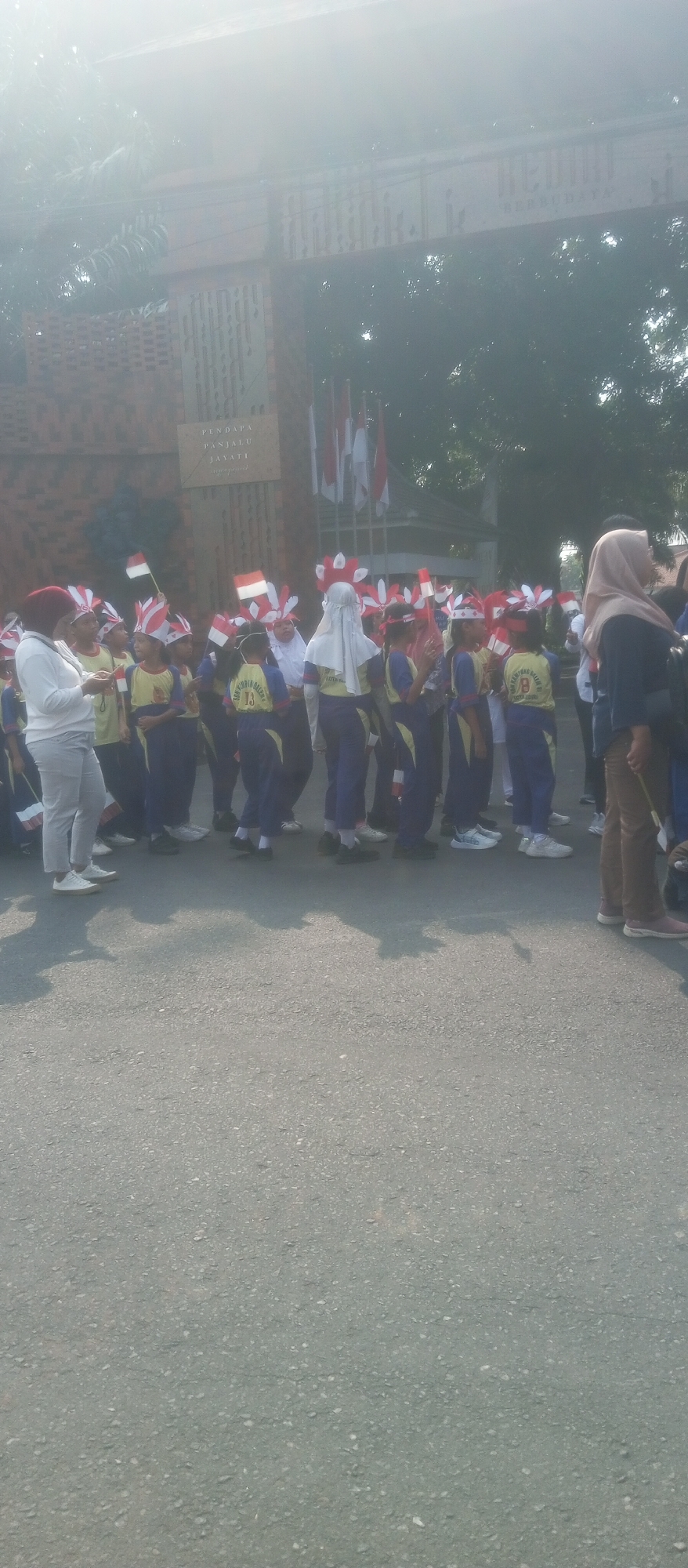 This image depicts an outdoor scene featuring a group of children, likely students, participating in a parade or event. Many of the children are wearing matching uniforms and decorative headbands with red and white elements, evoking national colors. Some children are holding small Indonesian flags. They are lined up on an asphalt road, facing towards the left of the frame. Behind them, a brick structure with decorative elements and text is visible, possibly a building or monument. Indonesian flags are also displayed prominently on poles above the brick structure. The overall atmosphere suggests a community event or celebration, possibly related to Indonesian Independence Day. The image is taken from a medium distance, focusing on the group of children and the background elements.